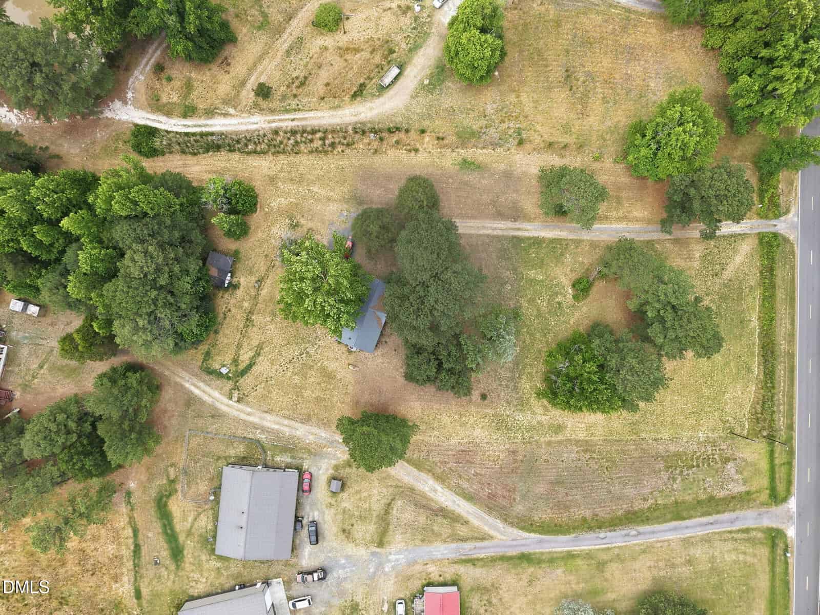 Aerial view of a rural property with a house surrounded by trees, dirt paths, a few parked vehicles, and adjacent roads. Nearby buildings and grassy areas are visible, with vegetation scattered throughout the landscape.