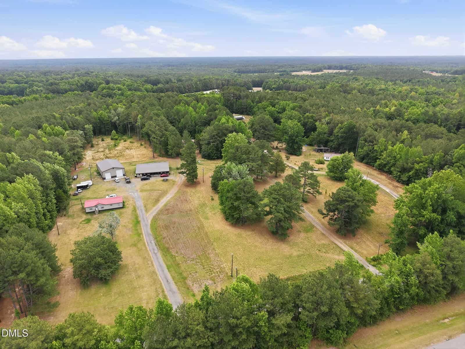 Aerial view of a rural property with several buildings, open grassy areas, trees, and winding driveways, surrounded by dense forest under a partly cloudy sky.
