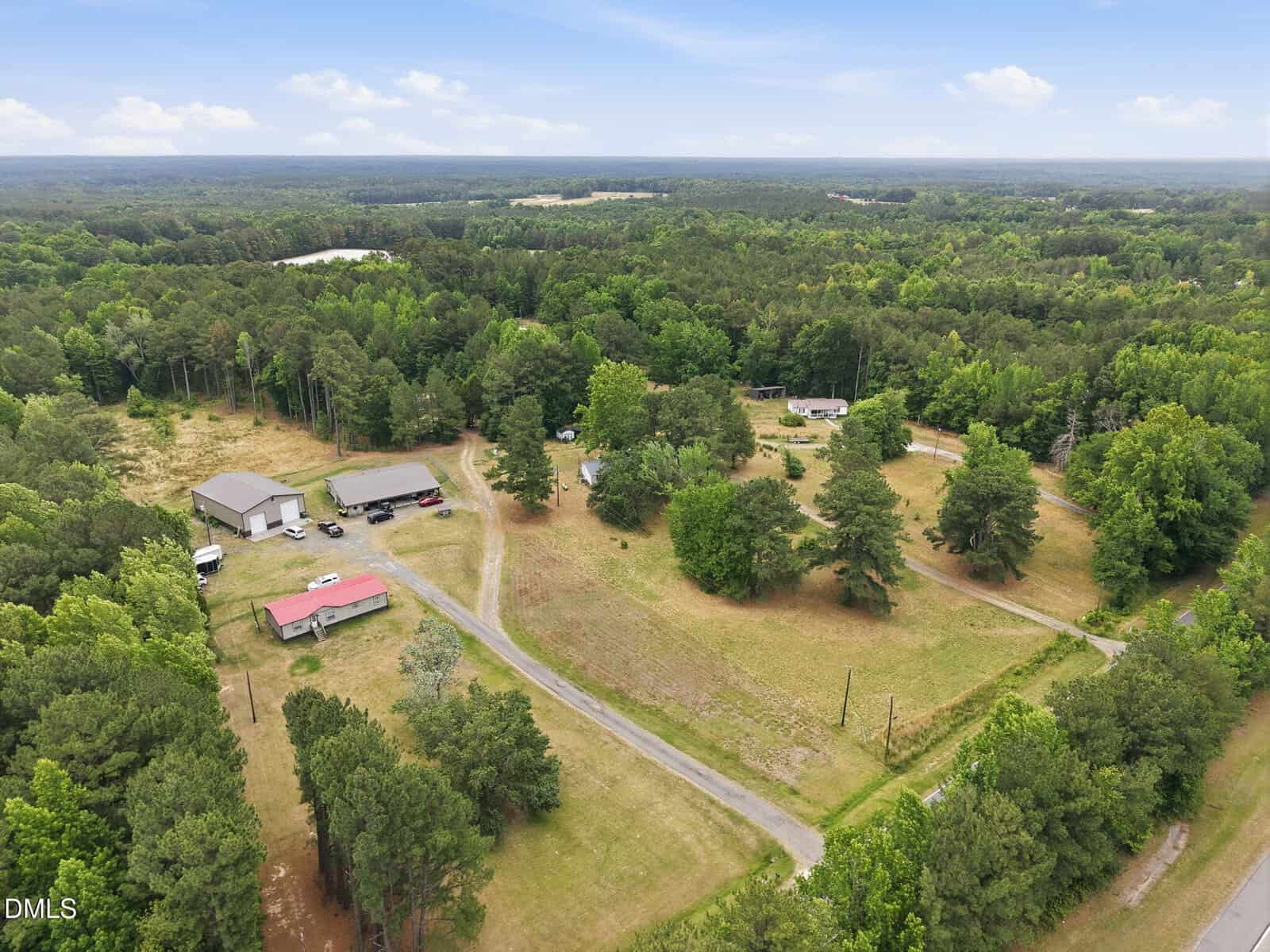 Aerial view of a rural property with multiple buildings, open grassy areas, scattered trees, and surrounding forest under a partly cloudy sky. A main road runs through the property.
