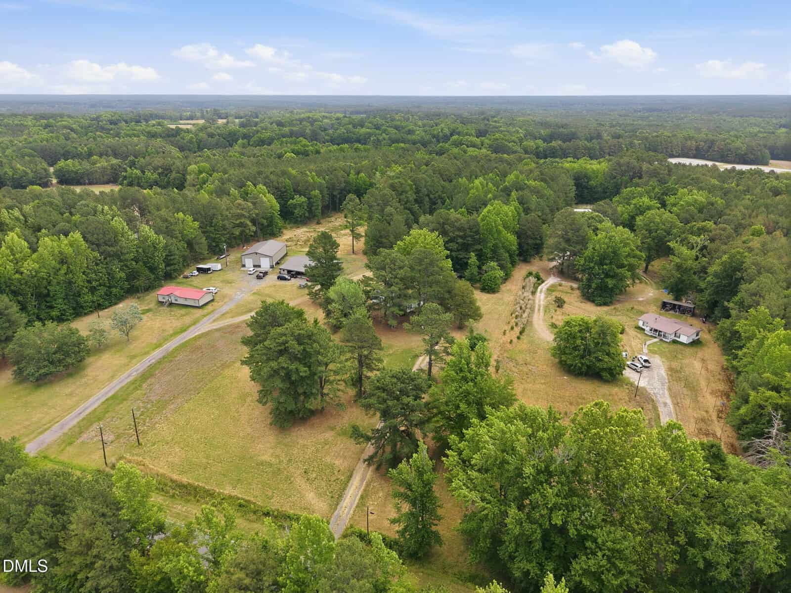 Aerial view of a rural property with two houses, surrounding trees, driveways, open grassy areas, and wooded landscape under a blue sky with clouds.