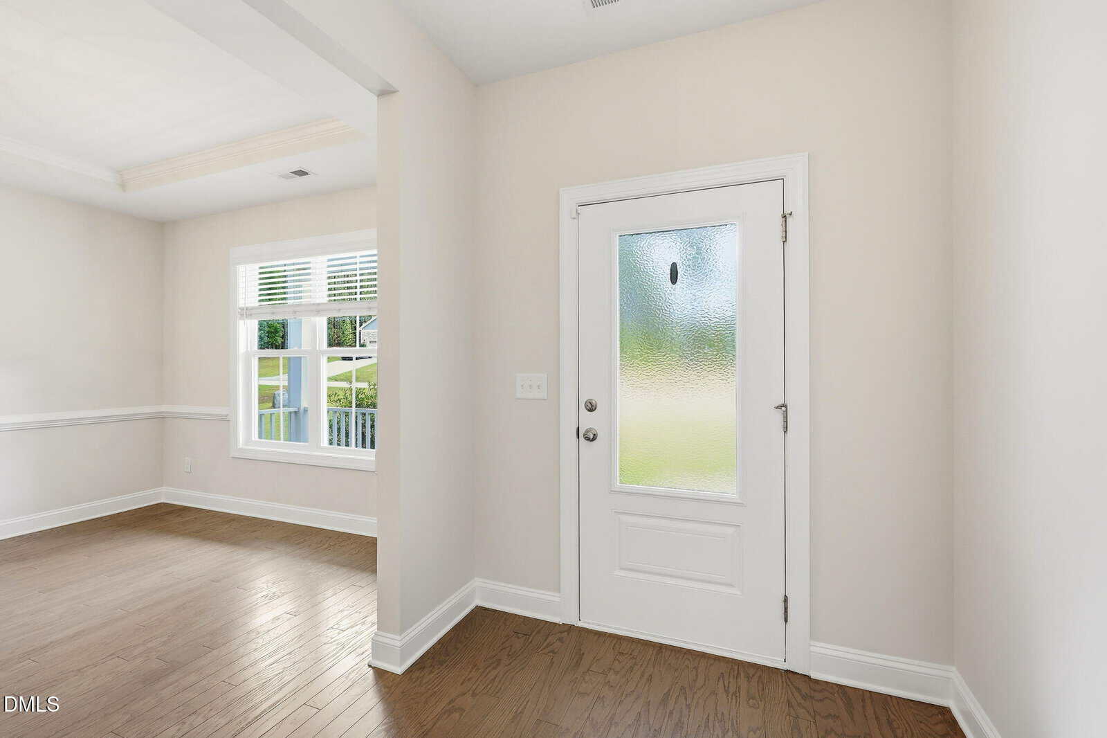A bright entryway with a white front door featuring a frosted glass panel, light-colored walls, and hardwood floors. A room with large windows and white trim is visible to the left.