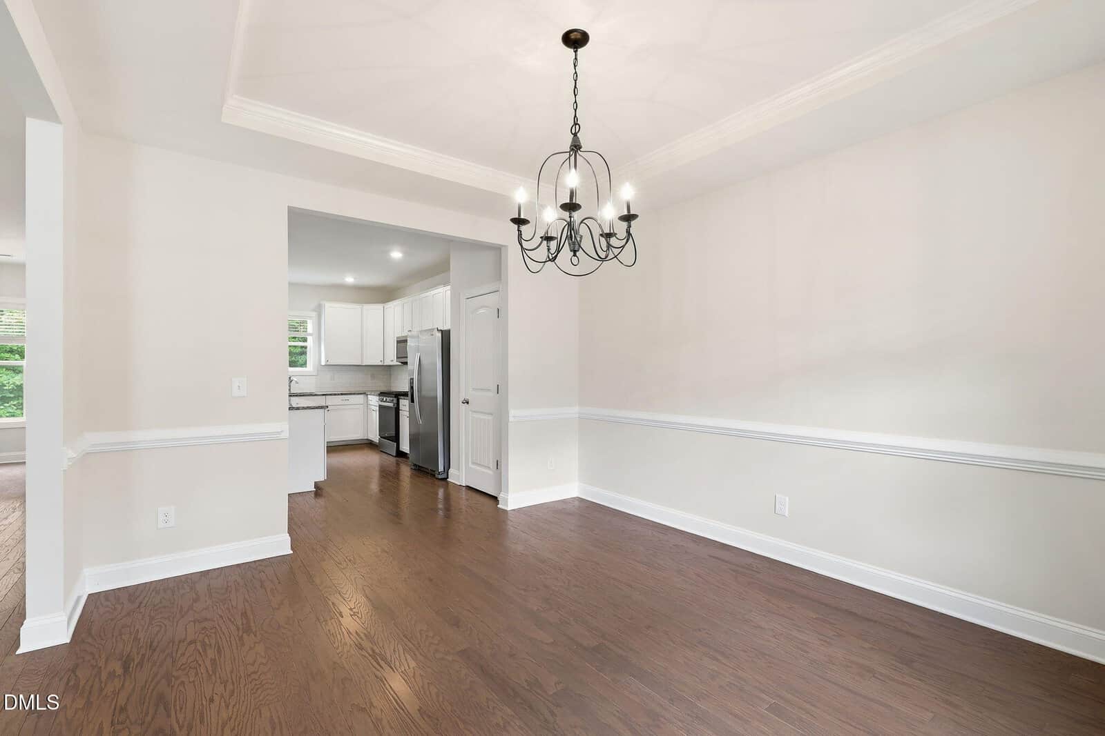 Bright dining room with light-colored walls, crown molding, and dark wood floors, featuring a black chandelier. The room opens into a modern kitchen with white cabinets and stainless steel appliances.