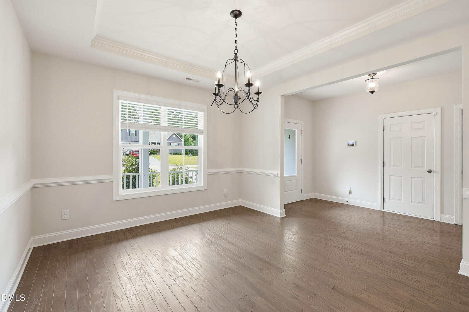 Bright, empty room with hardwood floors, white walls, a large window with blinds, a decorative ceiling light fixture, and a doorway leading to another area with two closed doors.