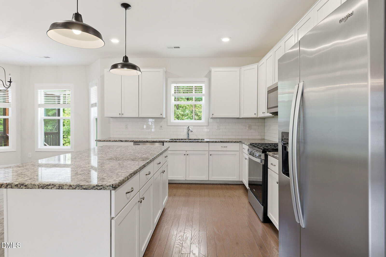 Modern kitchen with white cabinets, granite countertops, stainless steel appliances, two pendant lights, hardwood floors, a large island, and windows letting in natural light.