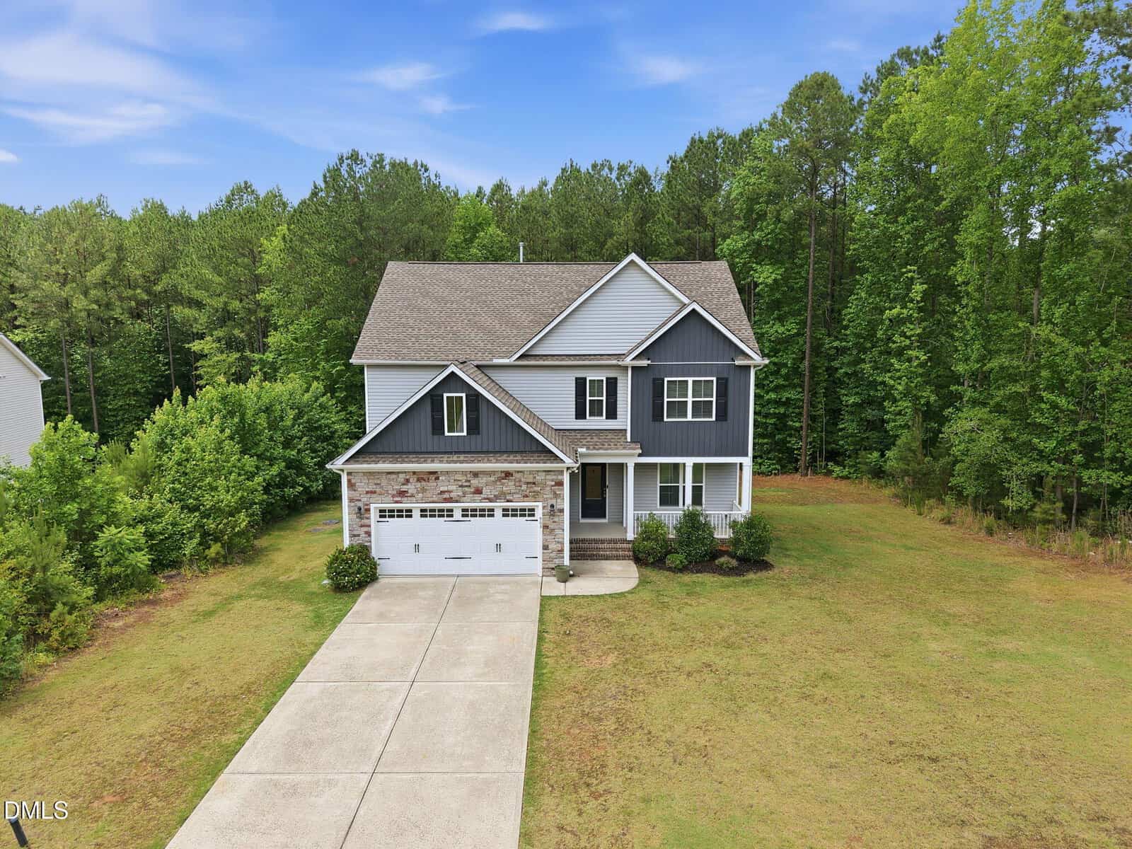 Two-story house with blue and gray siding, white trim, and attached two-car garage. Surrounded by a large lawn and backed by dense green trees under a blue sky. Concrete driveway leads to the garage.