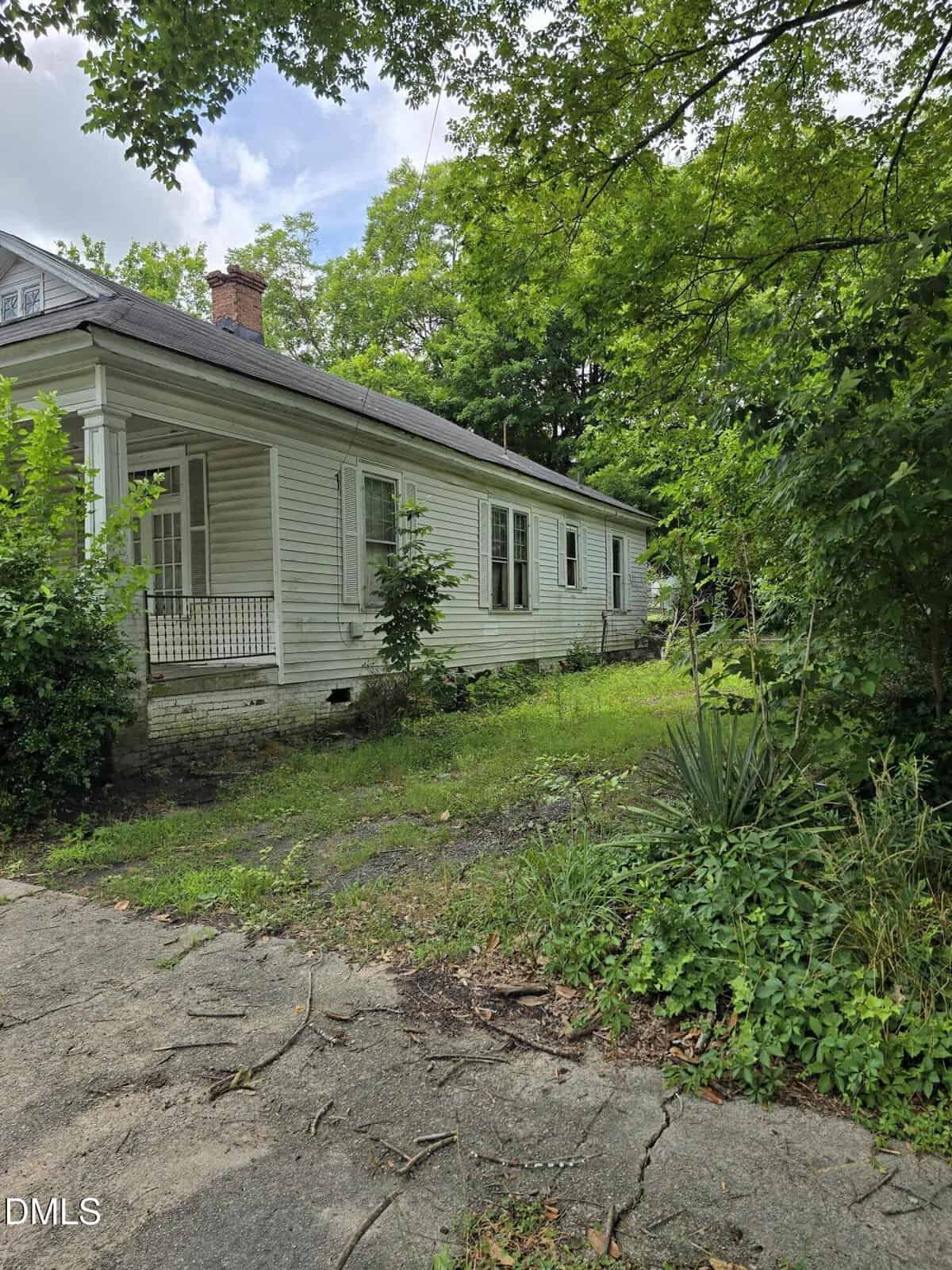 A white, single-story house with a small porch and multiple windows is surrounded by overgrown vegetation and trees. The concrete driveway shows cracks and patches of grass.