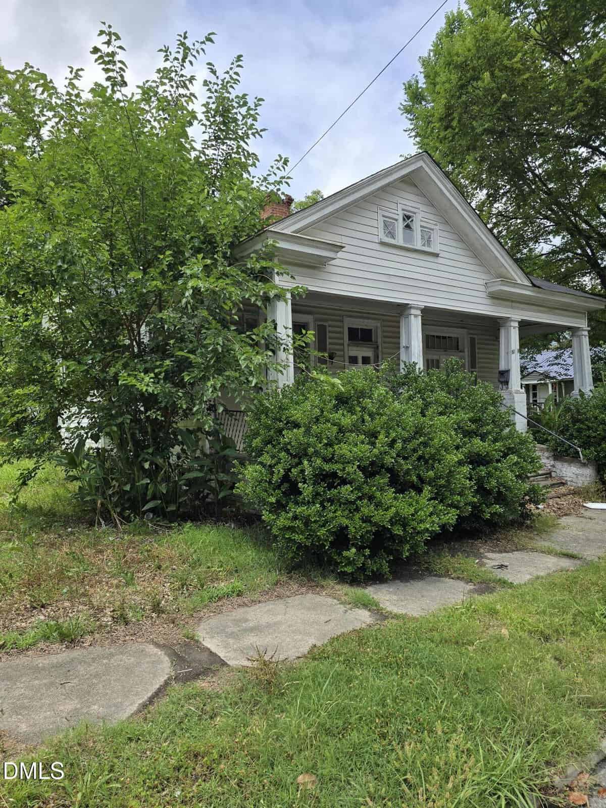 A white house with a front porch and columns is partly hidden by overgrown bushes and trees. The yard has uneven grass, and a stone walkway leads to the porch steps.