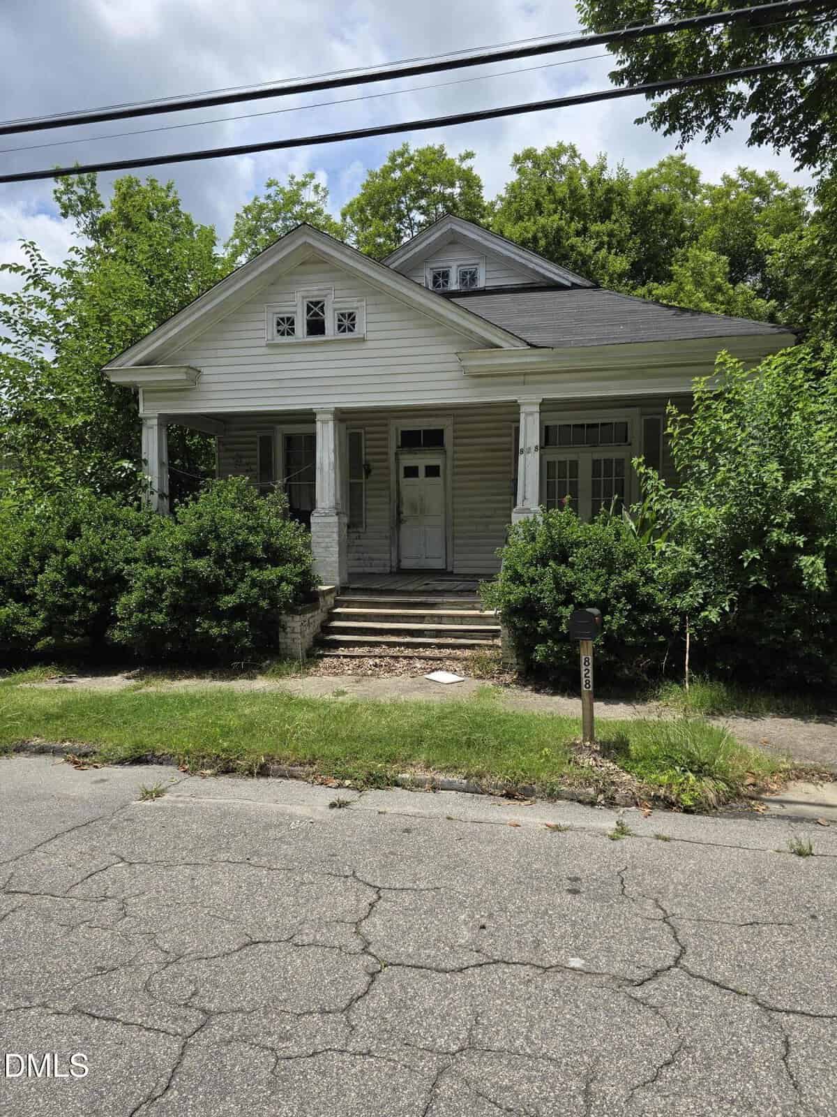 An old, white house with a front porch and pillars sits at 828 Nicholas Street in Henderson, surrounded by overgrown bushes and trees. The roof and steps show signs of wear, while a cracked sidewalk and street are visible in the foreground.