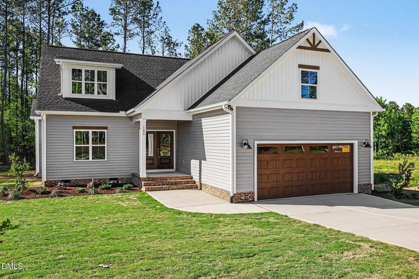 Modern two-story house at 105 Spring Grove Drive, Oxford, featuring light gray siding, a covered front porch, and a wooden double garage door. The neatly landscaped lawn is framed by trees under a clear blue sky.