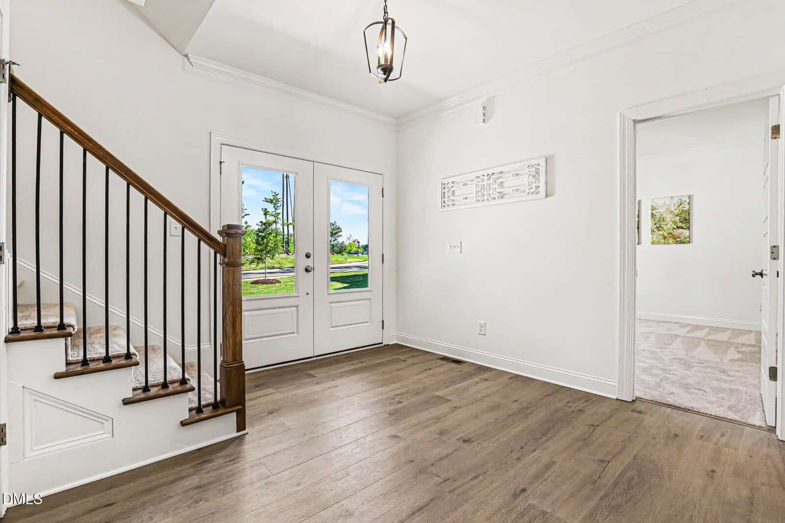 Bright entryway with wooden floors, white walls, and a staircase with dark railings. A glass-paneled double door leads outside; a decorative piece hangs above a light switch, and an open door reveals part of a carpeted room.