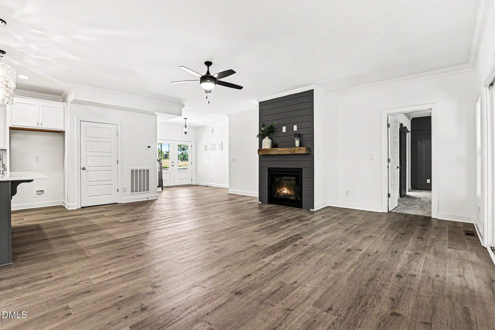 Open-concept living area with wood flooring, white walls, a black fireplace with a mantel, ceiling fan, open kitchen space, multiple doors, and natural light from windows in the background.