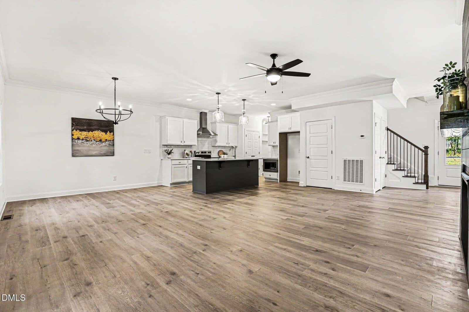 Open-concept modern kitchen and living area with light wood floors, white cabinets, a black island, stainless steel appliances, pendant lights, chandelier, and staircase leading upstairs.