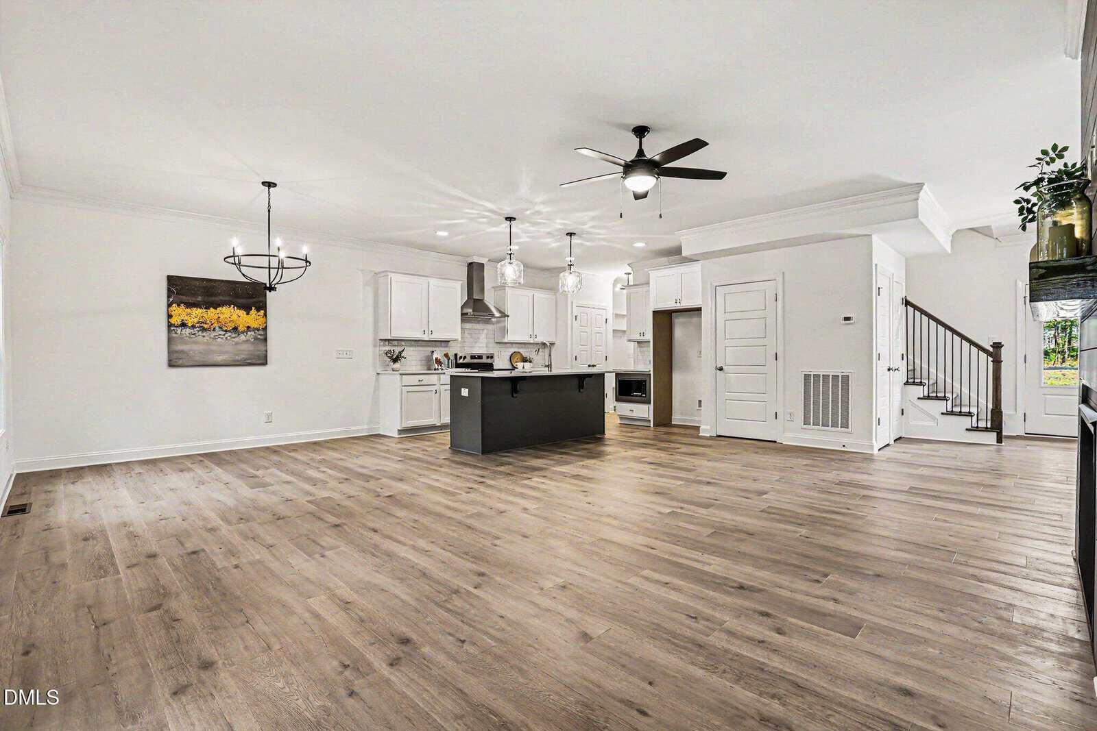 Open-concept kitchen and living area with light wood floors, white cabinets, a black island, modern light fixtures, ceiling fans, and a staircase leading upstairs. The space is bright and spacious.