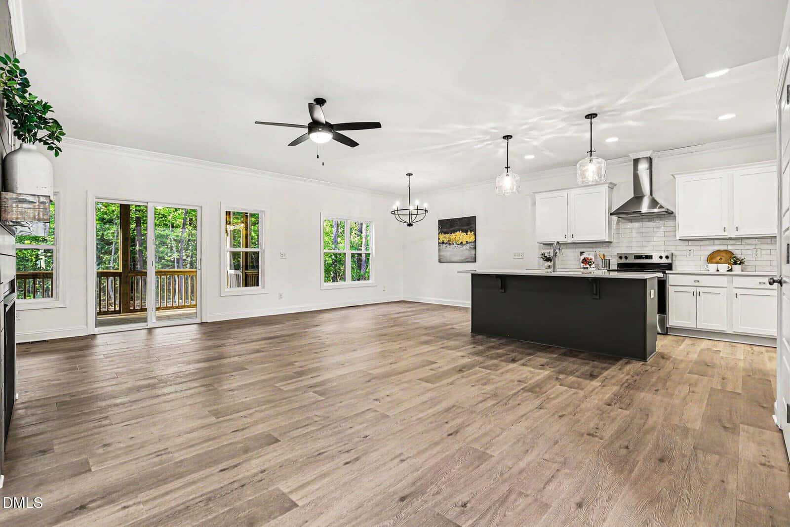 Spacious open-concept kitchen and living area with hardwood floors, white cabinets, a large island, ceiling fans, pendant lights, and large windows overlooking a wooded deck.