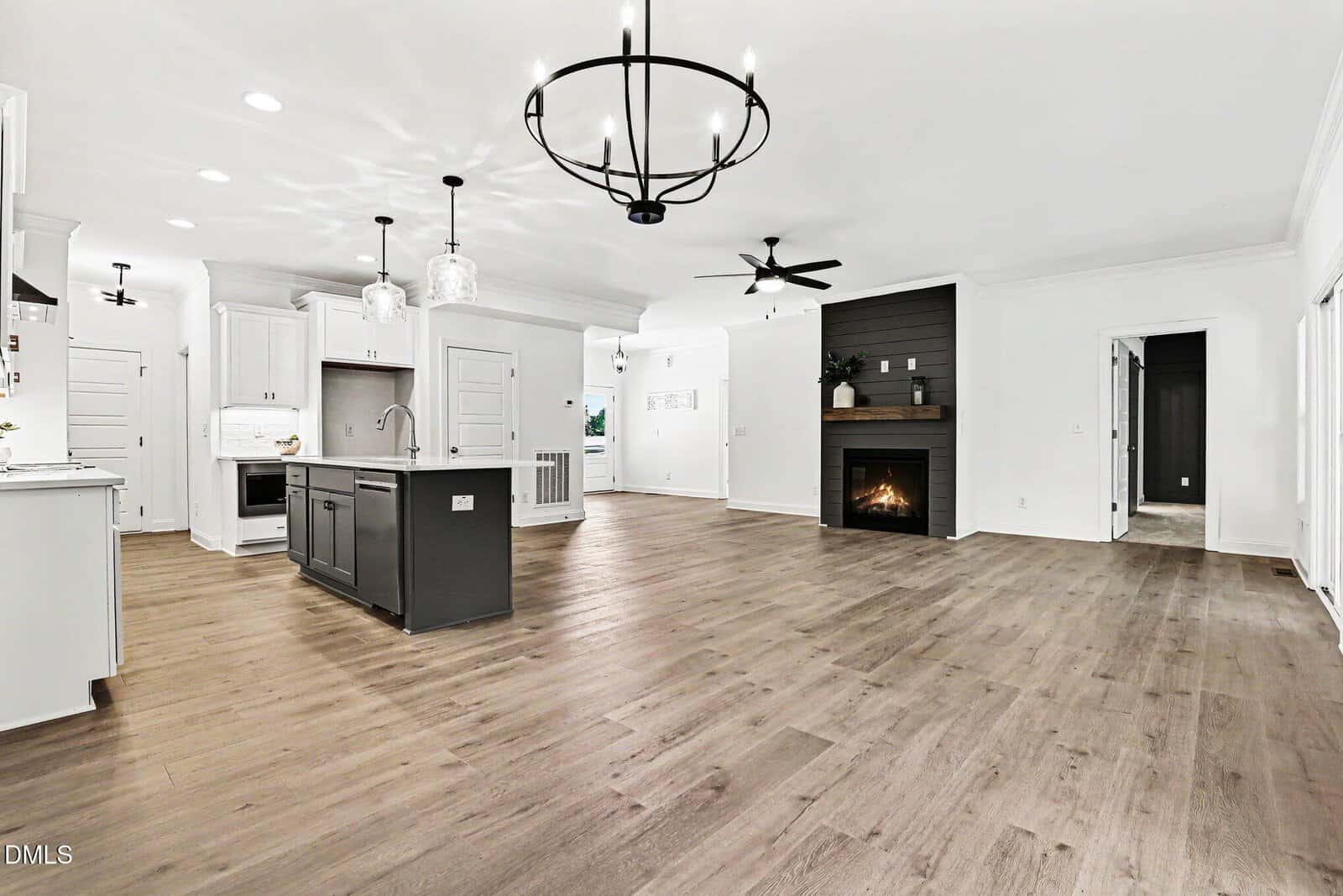 Modern open-concept living space with light wood floors, white kitchen cabinets, black island, two pendant lights, a fireplace with dark paneling, and large black chandeliers.