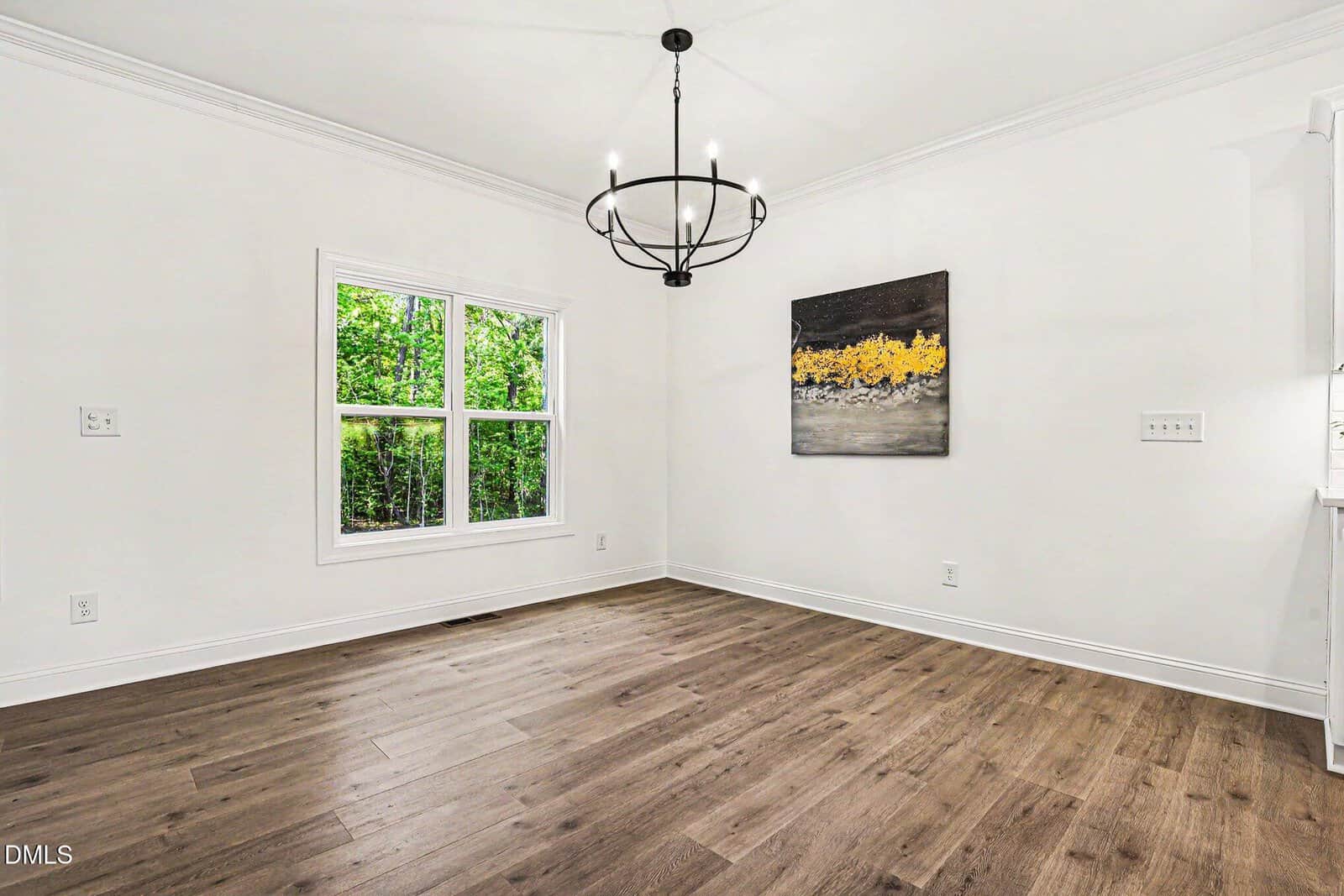 Bright, empty room with light walls, wood flooring, a large window showing greenery outside, a modern black chandelier, and a painting with yellow and gray tones on the wall.