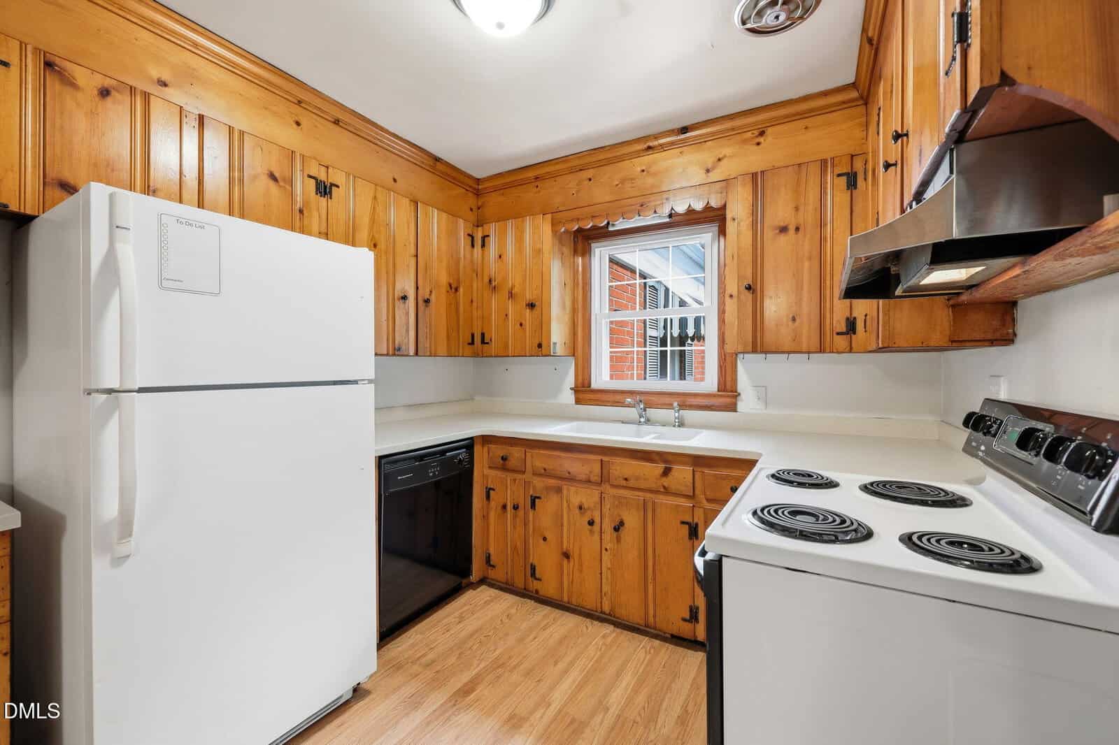 Cozy kitchen with wood-paneled cabinets, white countertops, white refrigerator, electric stove, black dishwasher, and a window above the sink letting in natural light.