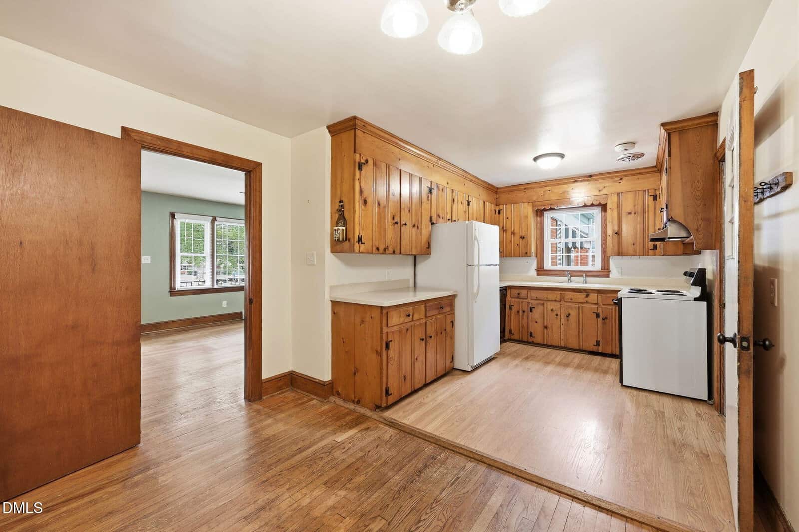 Kitchen with wood cabinets, white appliances, and wood floors. There’s a window above the sink, a door to another room, and a hanging light fixture on the ceiling. The room has a warm, rustic feel.