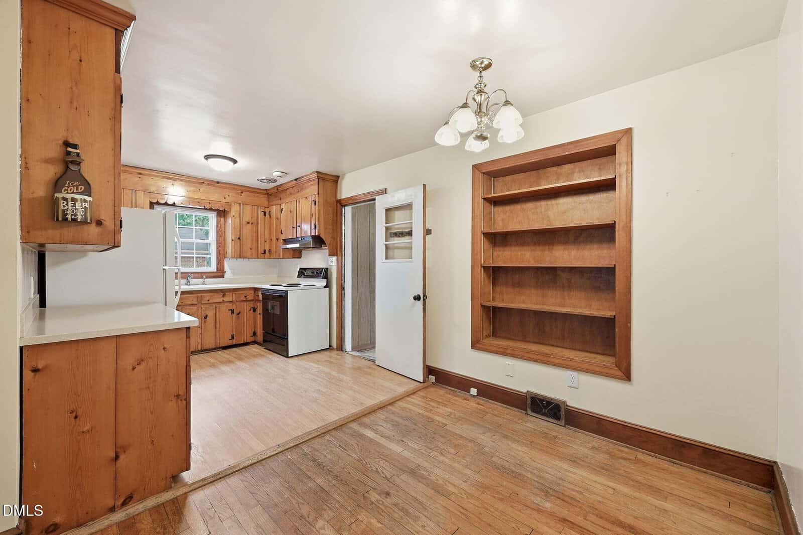 A kitchen with wood-paneled walls, white countertops, a stove, open shelving, and a small dining area with a built-in bookcase. Light wood flooring runs throughout the space.