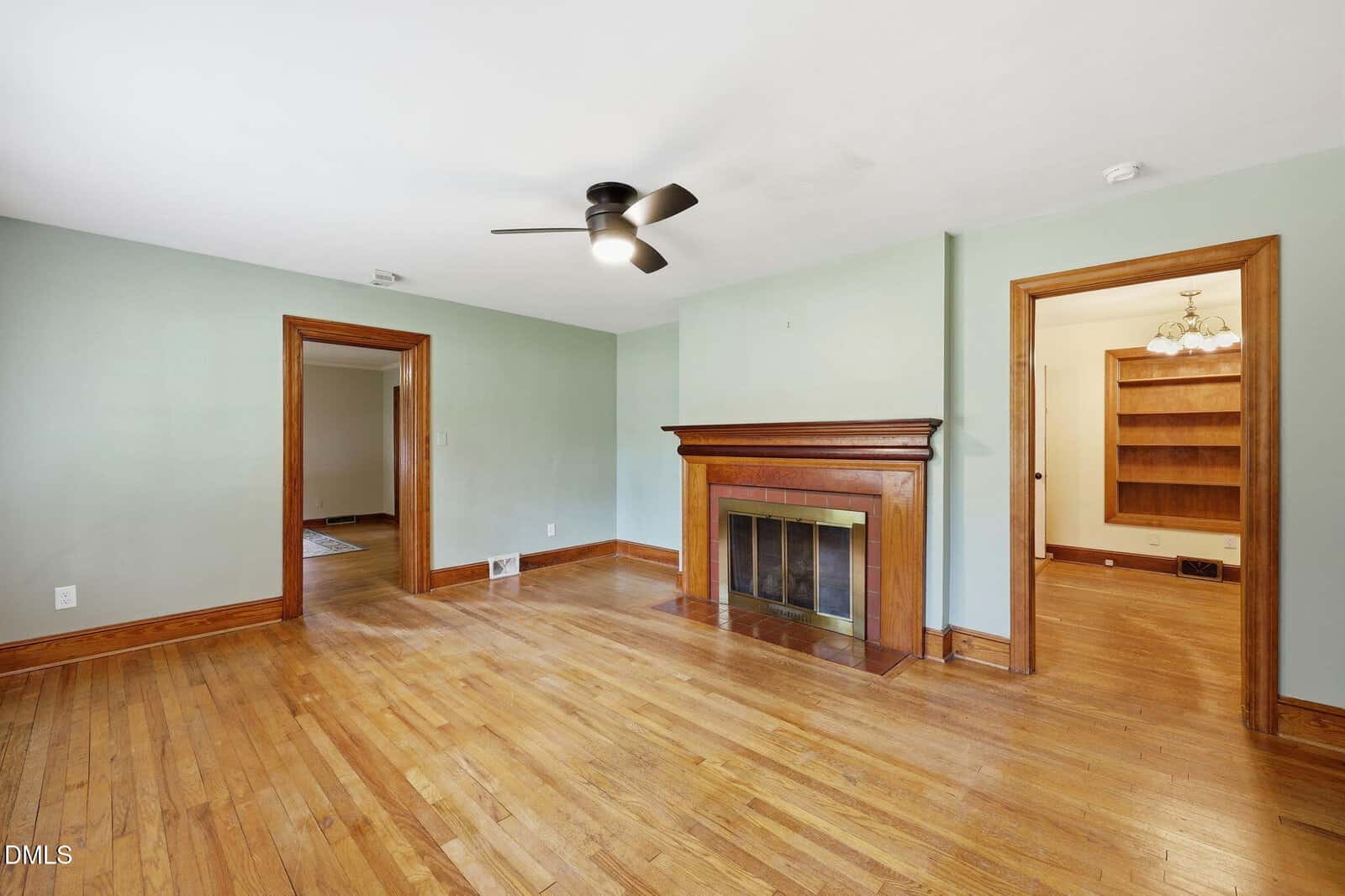 Spacious living room with light hardwood floors, a ceiling fan, a fireplace with a wooden mantle, pale green walls, and doorways leading to other rooms, one of which has built-in wooden shelves.