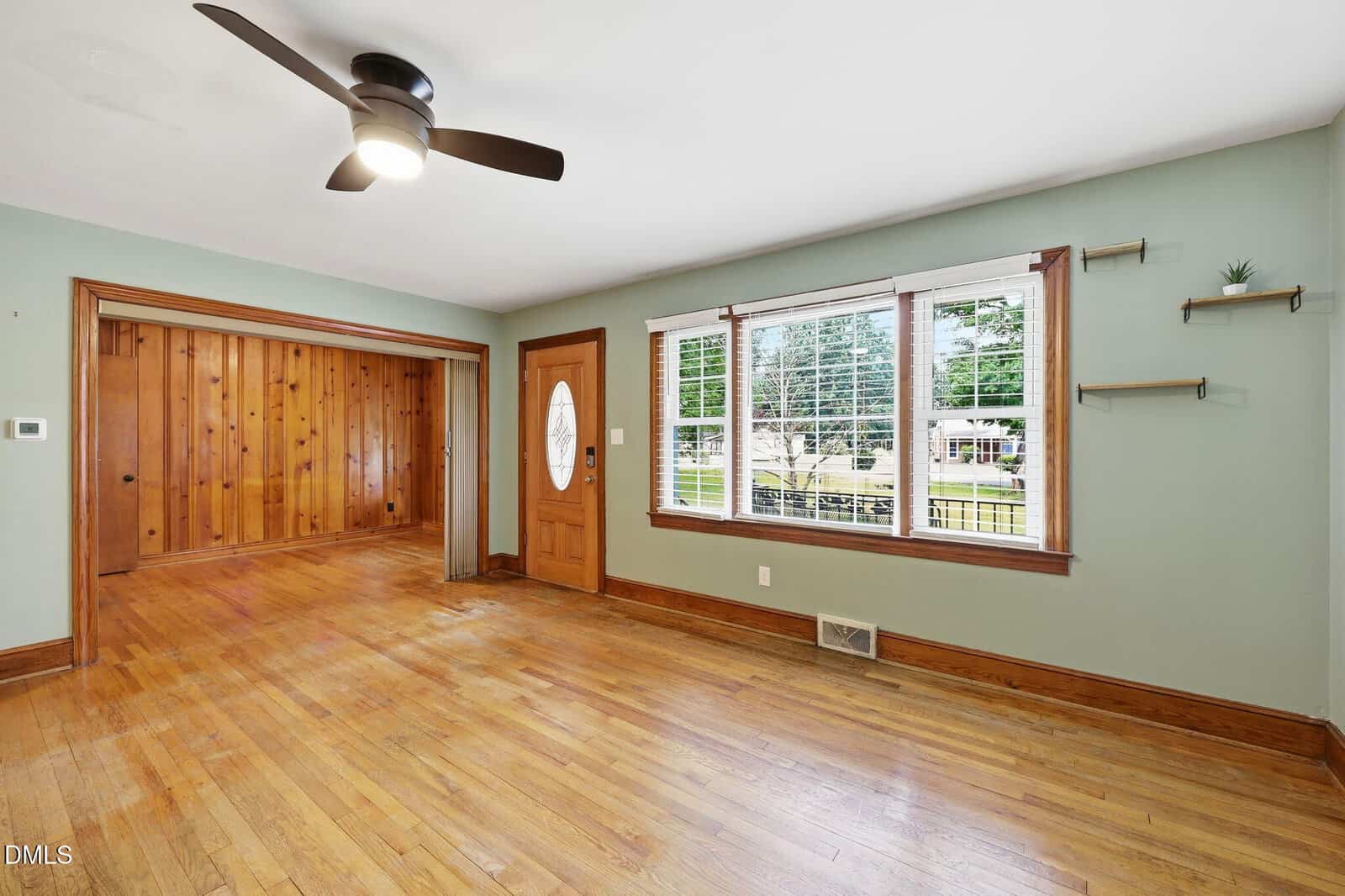 A bright living room with hardwood floors, light green walls, a wooden front door, large windows with white blinds, and an adjacent room with wood-paneled walls. Ceiling fan and floating shelves are also visible.
