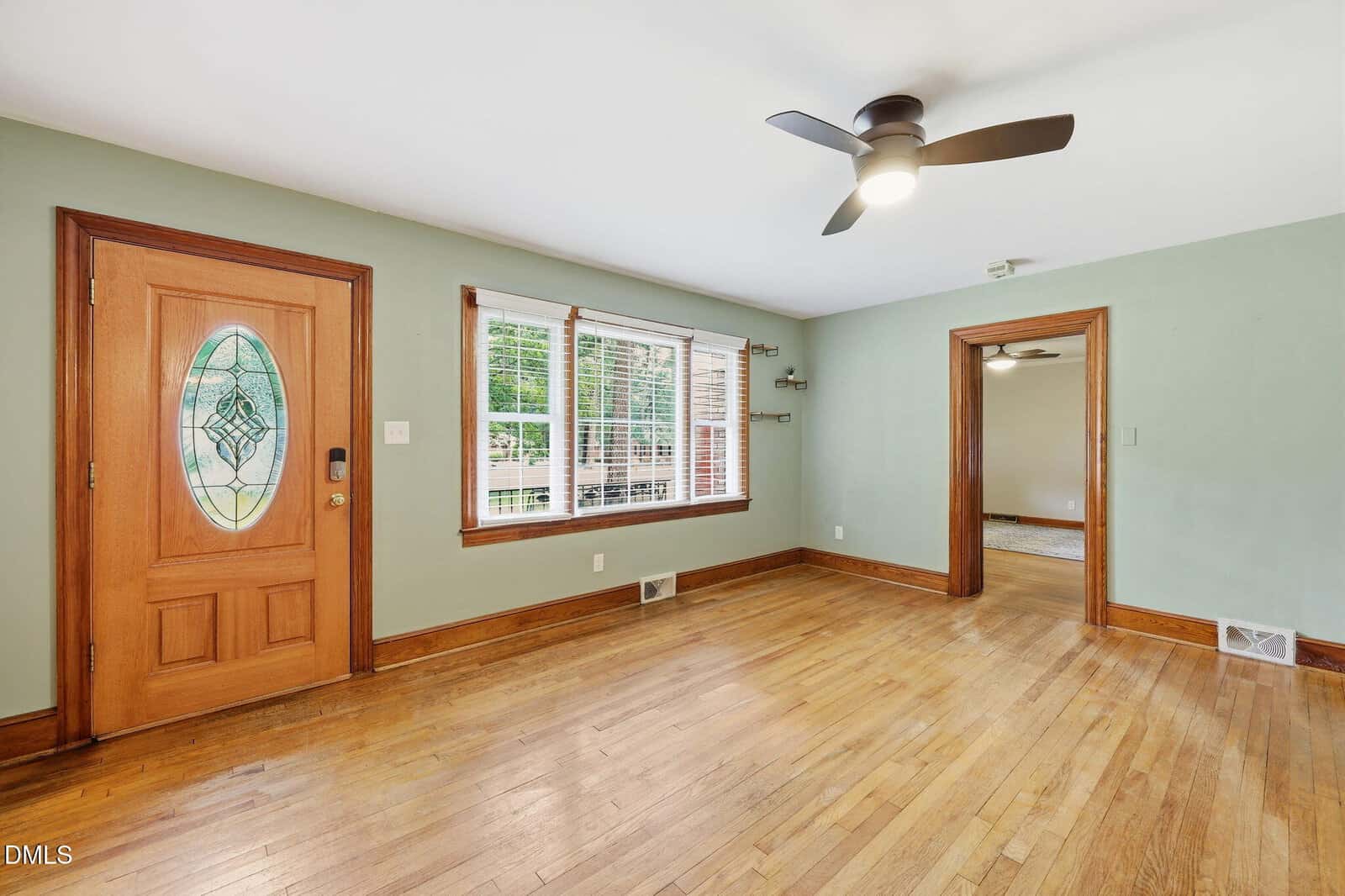 A bright, empty living room with light green walls, wood trim, a wooden front door with decorative glass, hardwood floors, large windows, and a ceiling fan. An open doorway leads to another room.