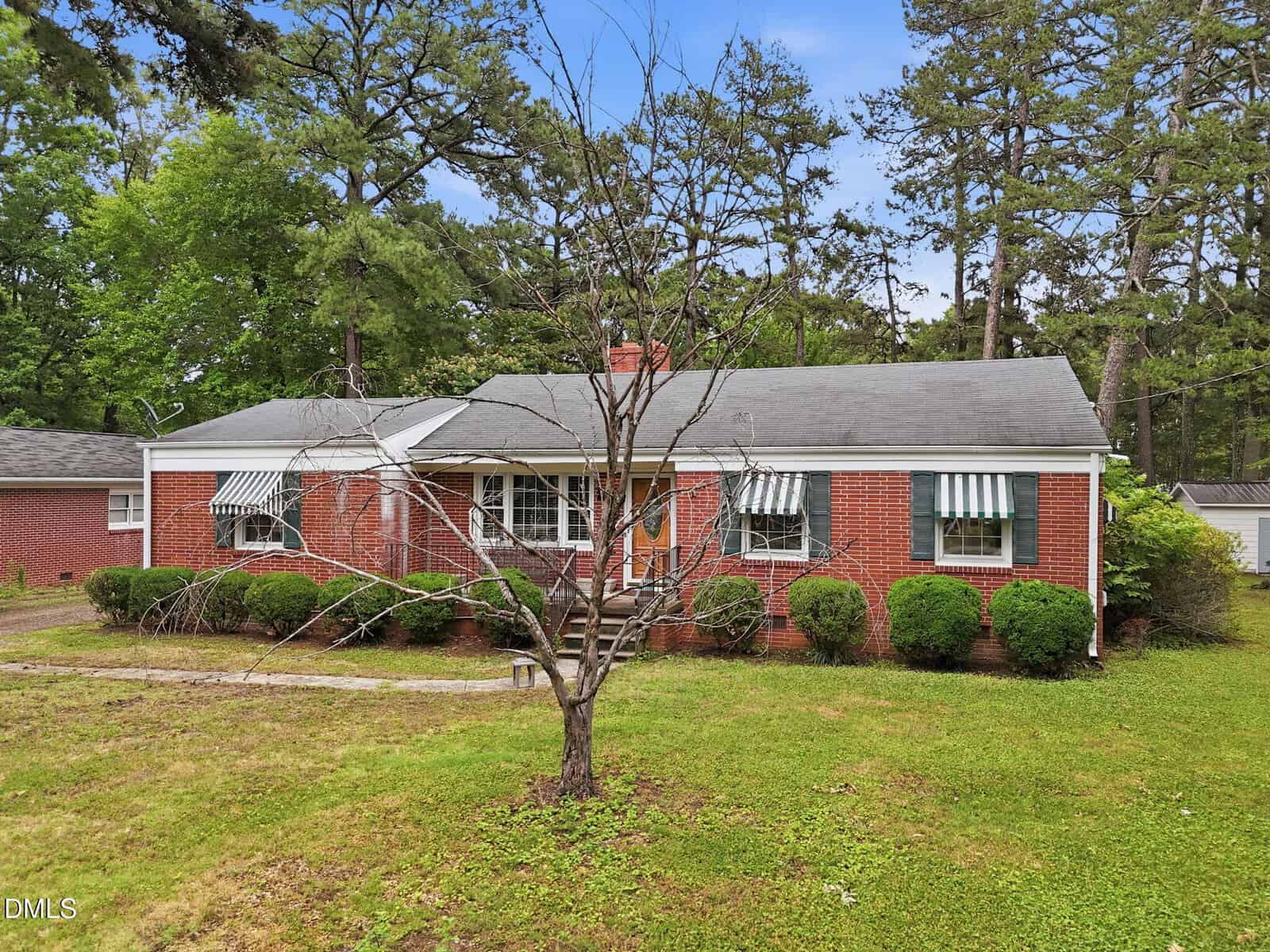 A single-story brick house in Oxford at 213 Pine Tree Road features green and white striped awnings, a front porch with steps, trimmed bushes, a leafless tree in the yard, and is surrounded by tall pine trees.