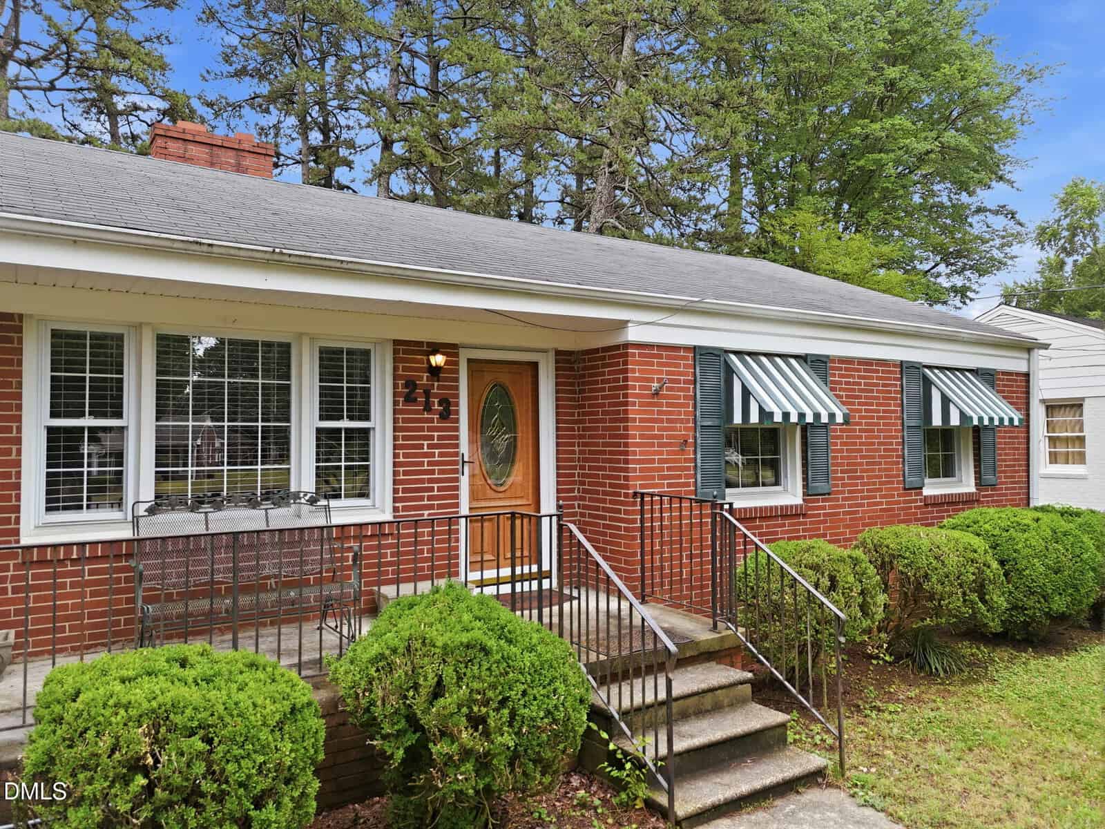Single-story brick house with white-trimmed windows, green-and-white striped awnings, a wooden front door with glass insert, metal railings, and neatly trimmed bushes lining the front walkway.