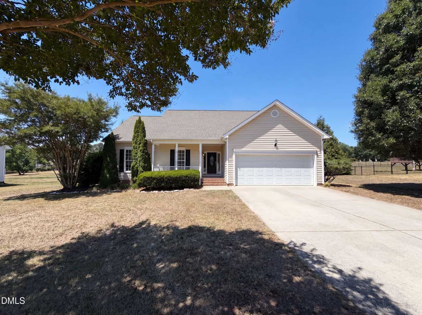 Charming single-story beige house at 20 Falling Leaf Drive, Youngsville, featuring a front porch, white garage, and driveway. The yard includes trees, bushes, and dry grass beneath a clear blue sky.