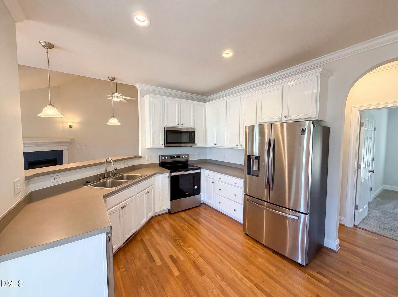 Modern kitchen with white cabinets, stainless steel appliances including a fridge, oven, microwave, and dishwasher, gray countertops, double sink, and hardwood floors. Room is well-lit with recessed and pendant lighting.