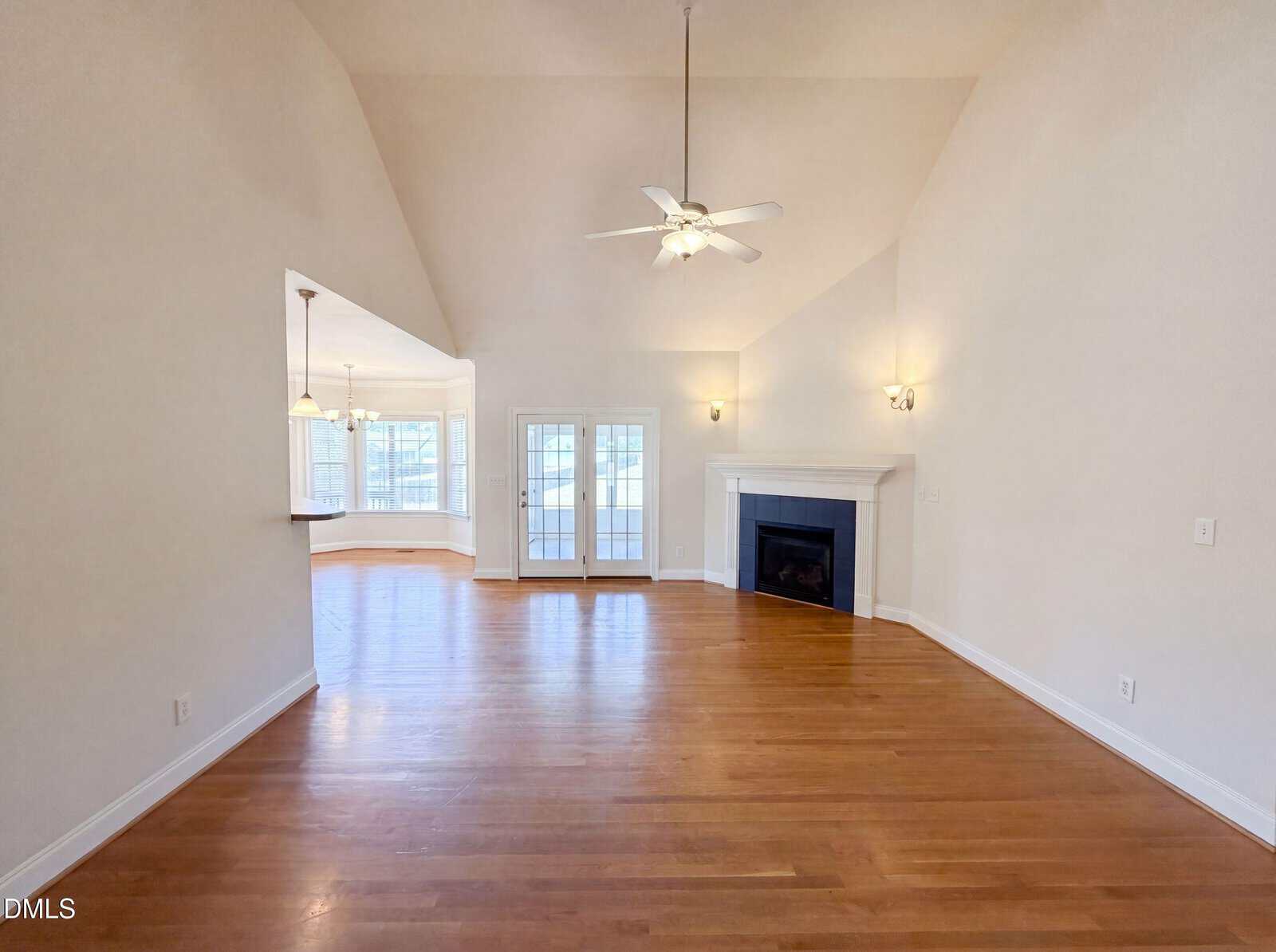Spacious, empty living room with hardwood floors, high vaulted ceiling, ceiling fans, a fireplace, and double glass doors leading to a bright sunroom or porch. The kitchen area is visible to the left.