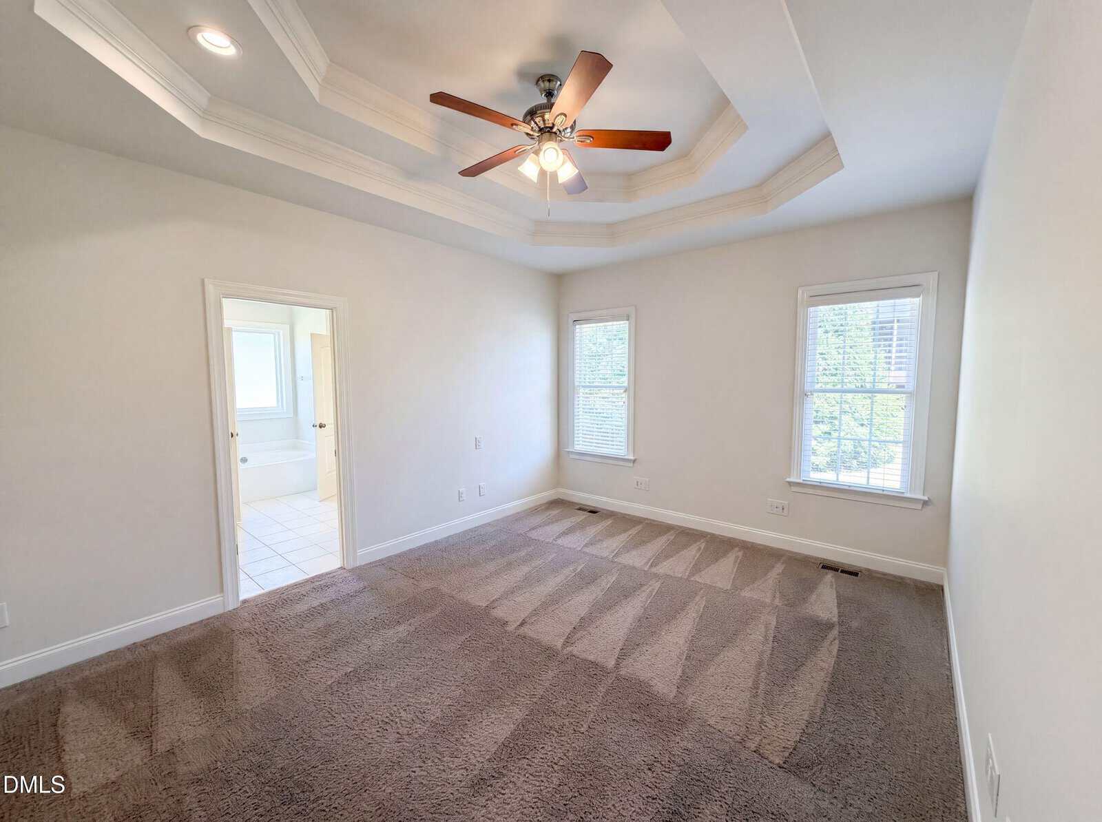 A bright, empty bedroom with tray ceiling, ceiling fan with lights, beige carpet, two windows, and an open doorway leading to a tiled bathroom.