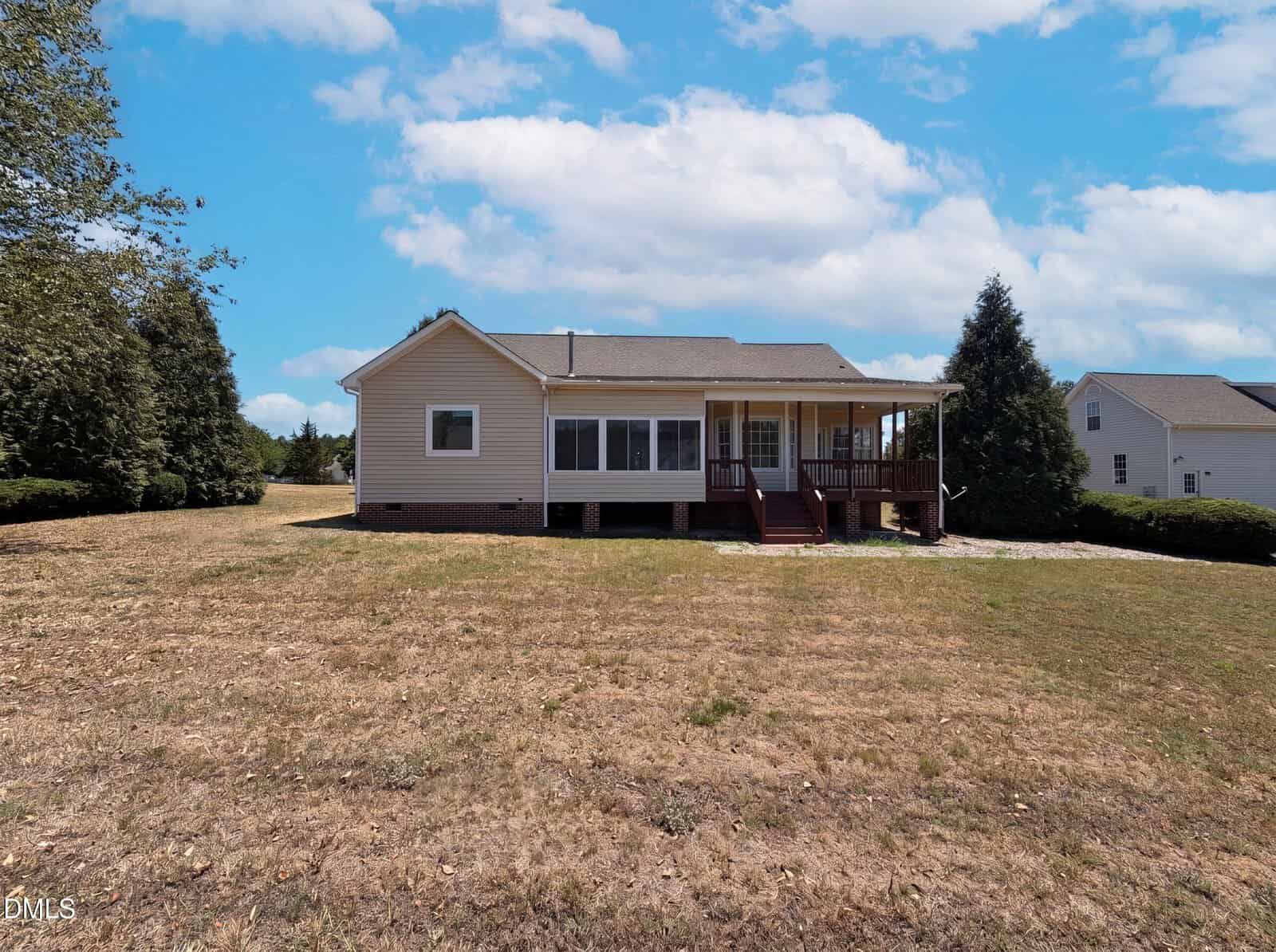 A single-story beige house with a covered porch and steps leading to a large, dry grassy yard. The sky is partly cloudy, and trees border the property in the background.