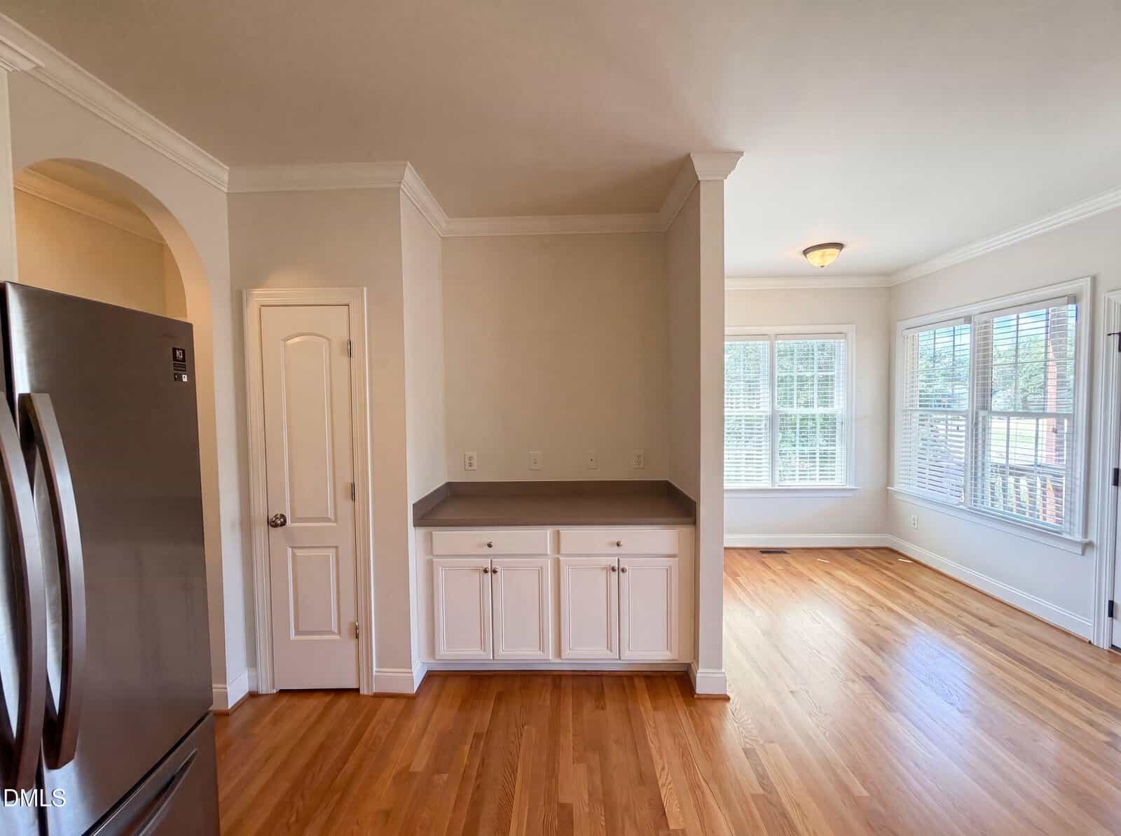 A kitchen with stainless steel fridge, white cabinets, and hardwood floors. Adjacent to a bright dining area with large windows and lots of natural light.