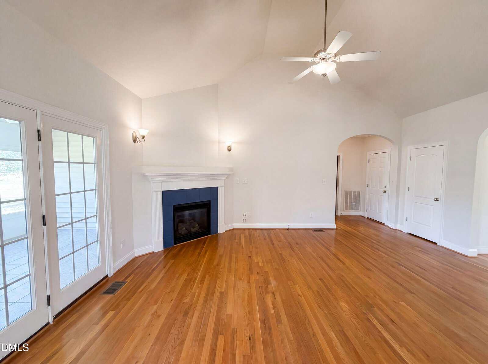 Spacious living room with hardwood floors, a fireplace, ceiling fan, vaulted ceiling, French doors to the left, and white walls with multiple doors on the right side.