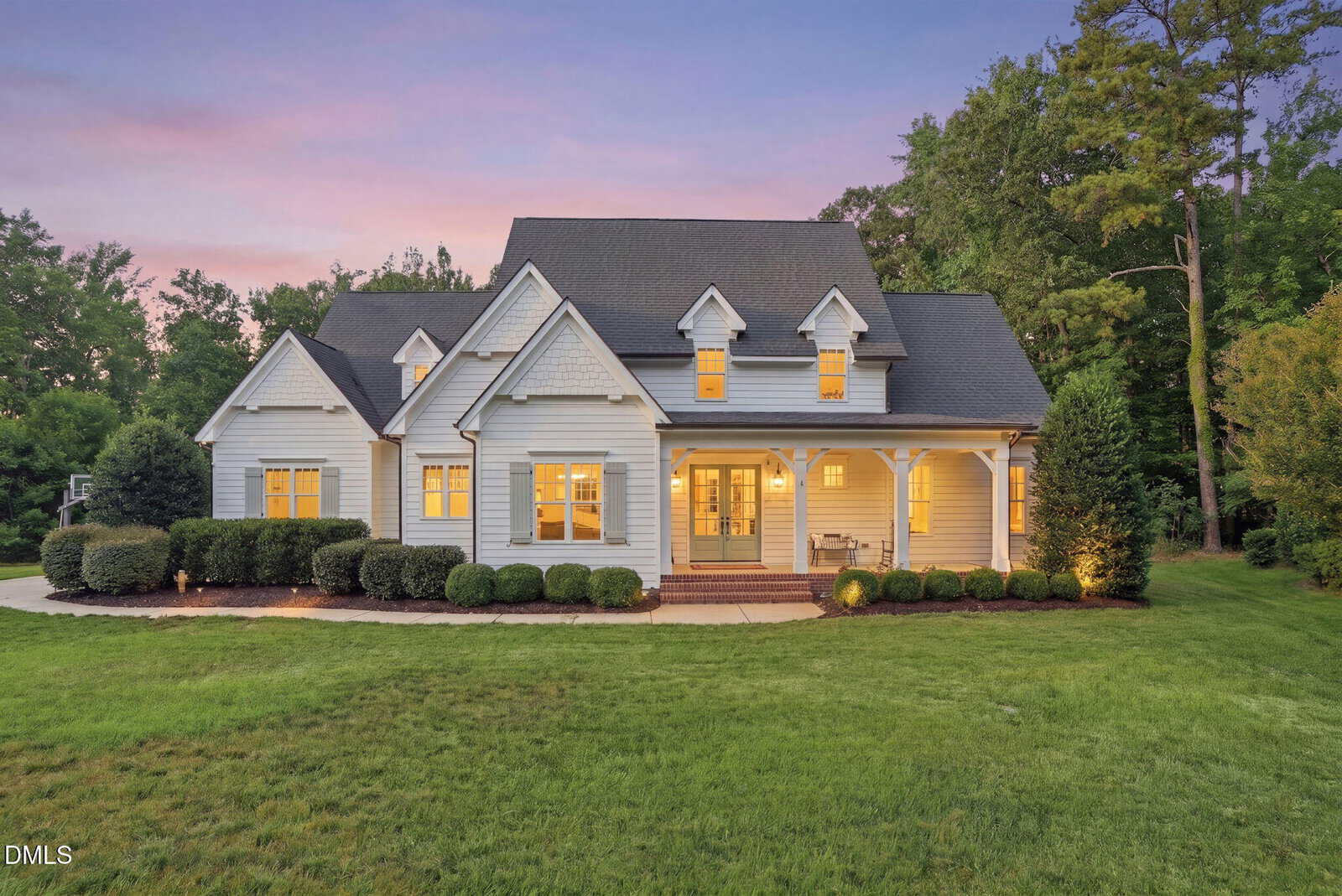 A charming white house with dormer windows at 335 Forest Bridge Road in Youngsville features a covered front porch and glowing interior lights, set on a large, well-manicured lawn with trees in the background at sunset.