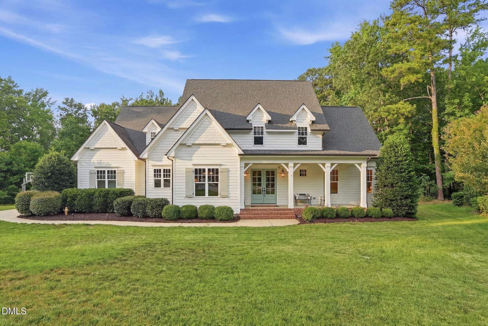 A two-story white house with a large front porch, dormer windows, and a gray roof, surrounded by green lawn, shrubs, and tall trees under a clear blue sky.