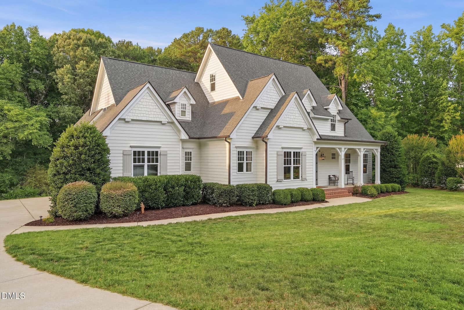 A white, two-story house with a gabled roof, dormer windows, and a covered front porch, surrounded by green grass, manicured bushes, and tall trees in the background.