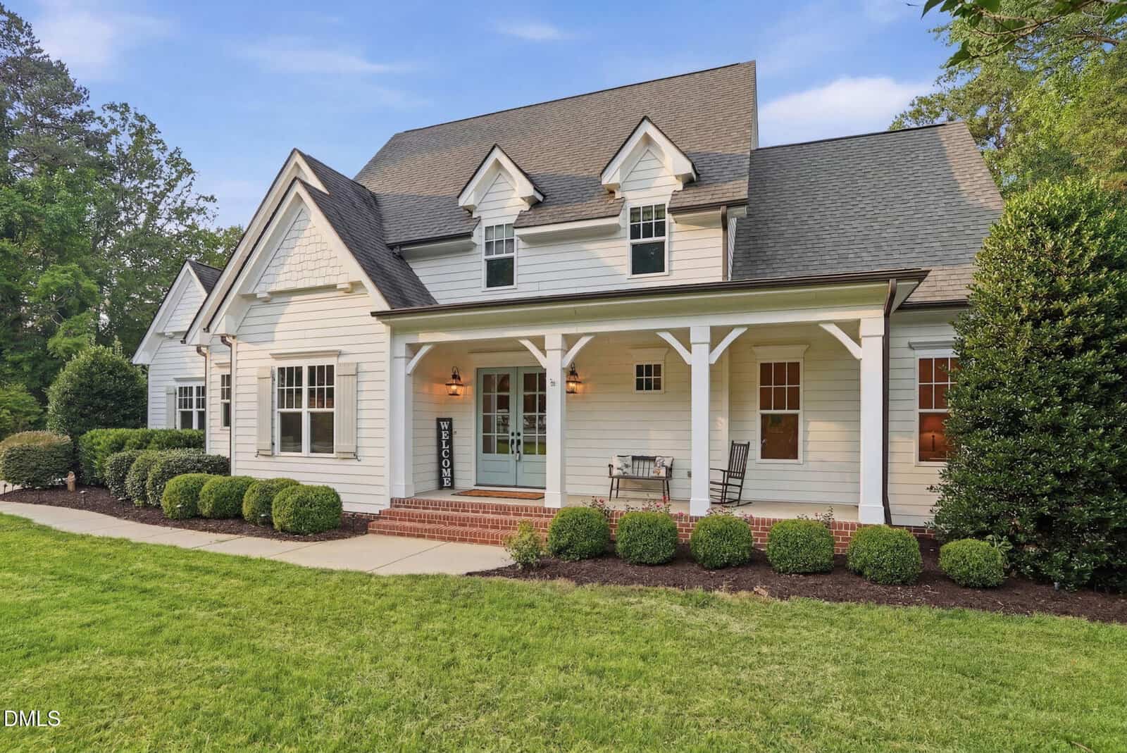 A two-story white house with a gabled roof, front porch with chairs, brick steps, tidy landscaping, trimmed bushes, and a lush green lawn under a blue sky.