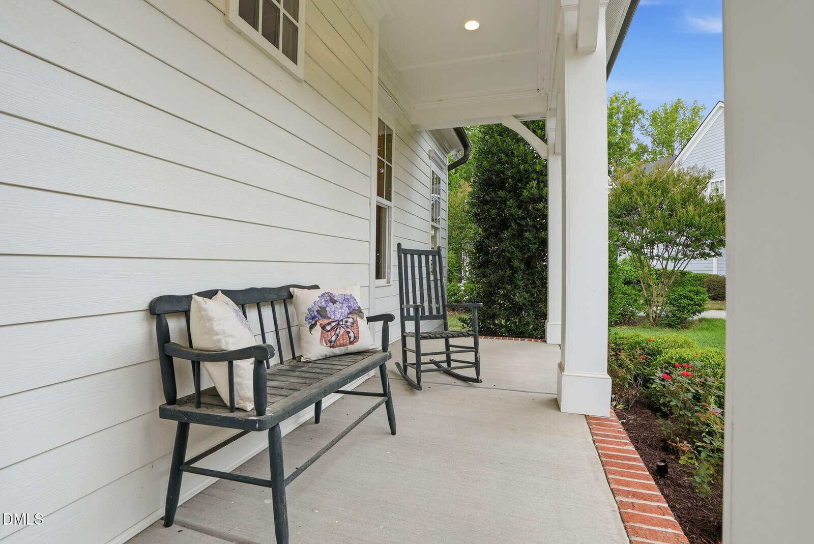 A cozy front porch with a black wooden bench, a cushion and pillow, and a black rocking chair on a concrete floor, next to white siding and a garden with green shrubs and flowering plants.