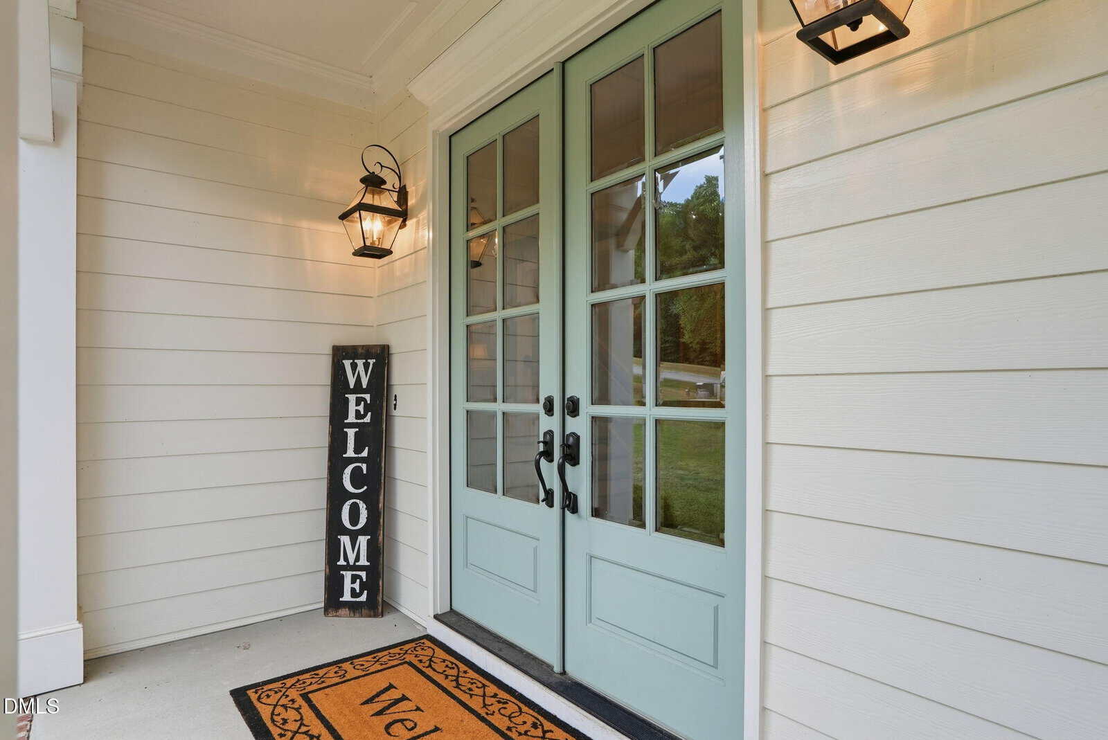 Light blue double doors with glass panes lead into a house. A welcome mat and a tall “Welcome” sign are by the entrance. Two black lantern-style lights are mounted on the white exterior wall.