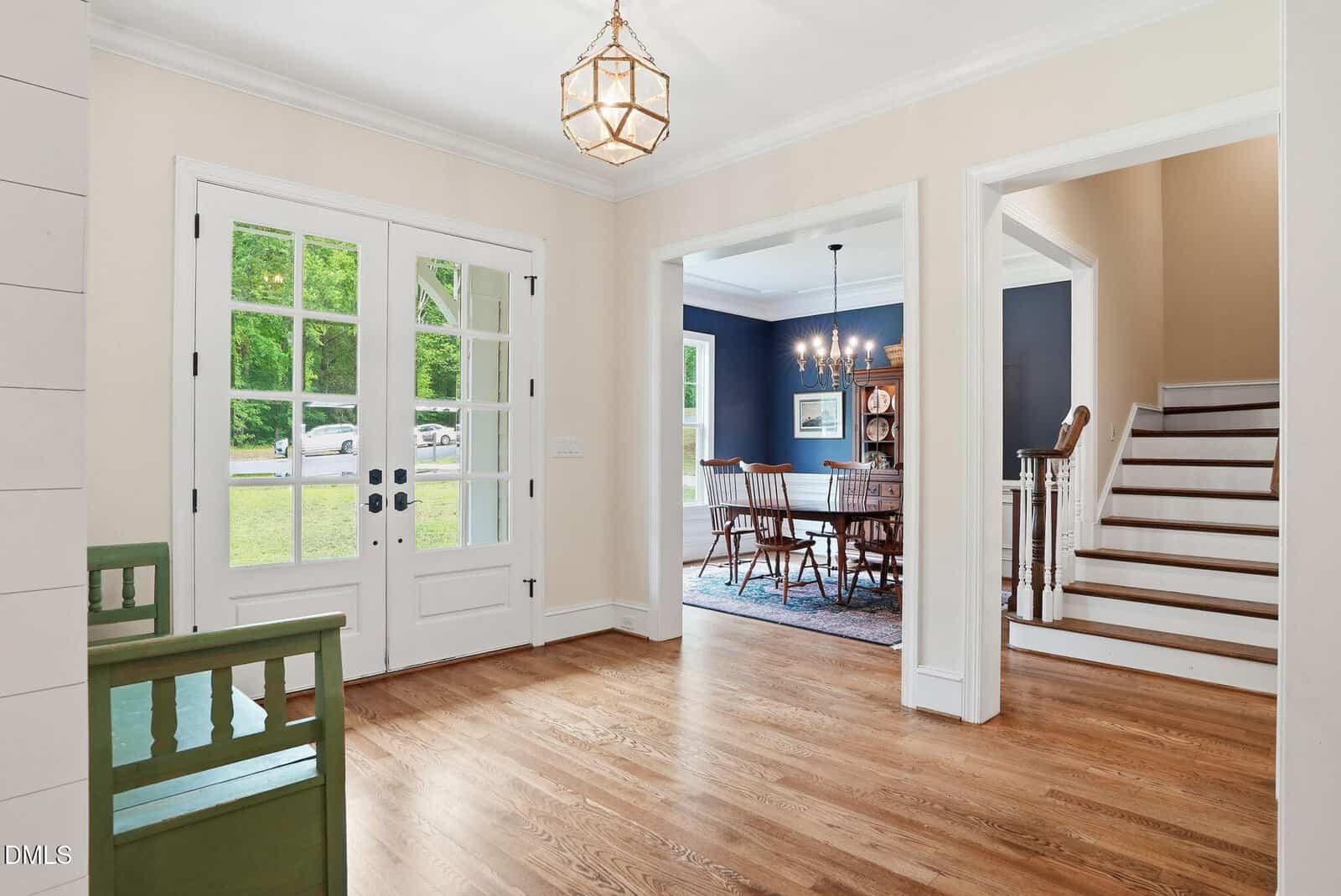Bright entryway with double glass doors, light wood floors, a view into a dining room with navy walls, chandelier, and wooden furniture; staircase leads upstairs on the right.