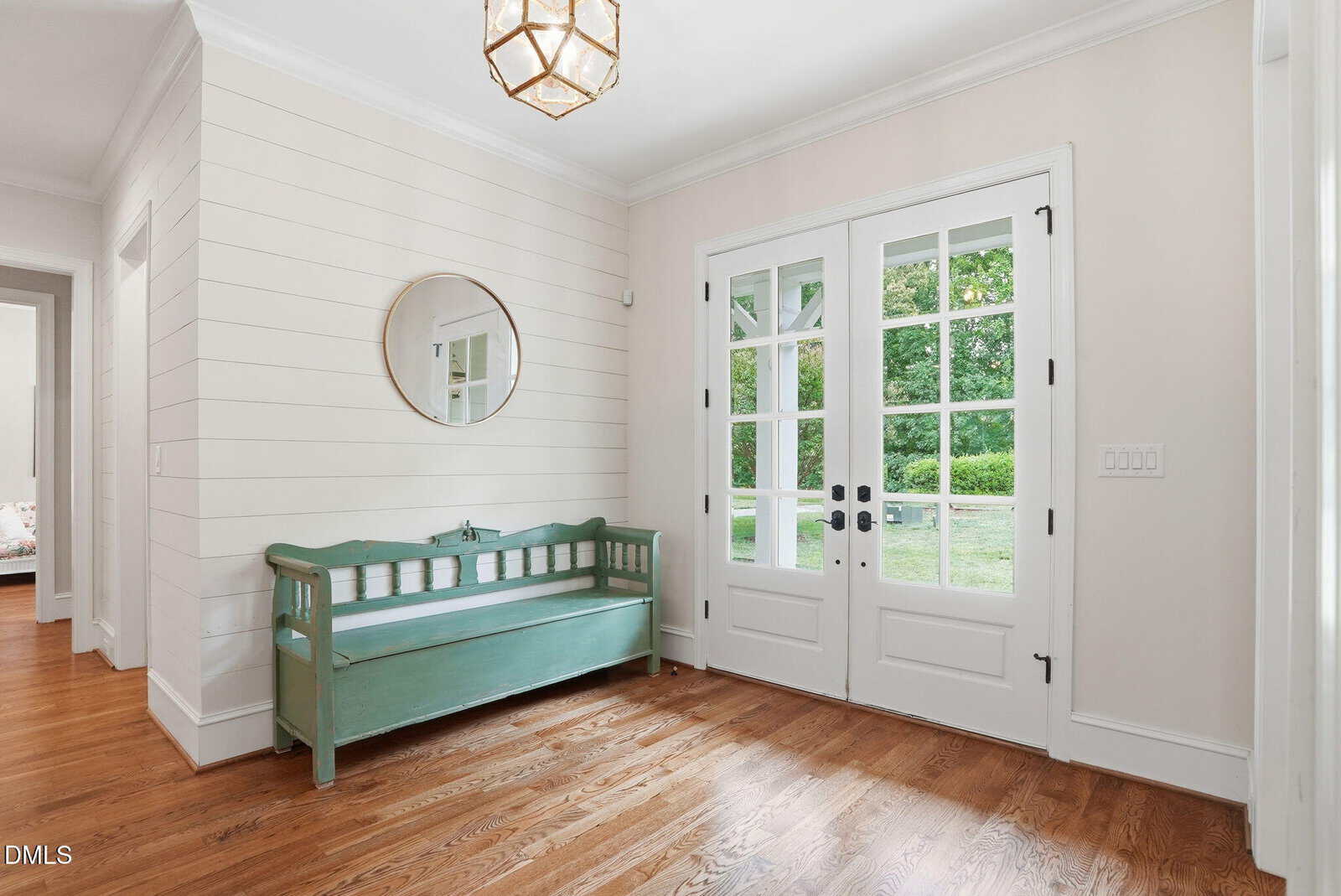 Bright entryway with wooden floors, a mint green bench, round mirror on a shiplap wall, double glass doors, and a geometric light fixture. Lush greenery is visible outside.