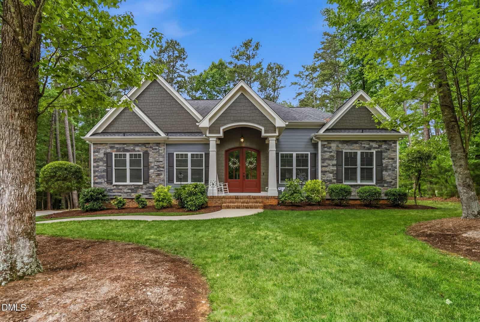 A single-story house at 3884 Whisperwood Court, Youngsville, featuring elegant stone and gray siding, a striking red double front door, arched windows, and a well-maintained lawn surrounded by trees and landscaping under a blue sky.