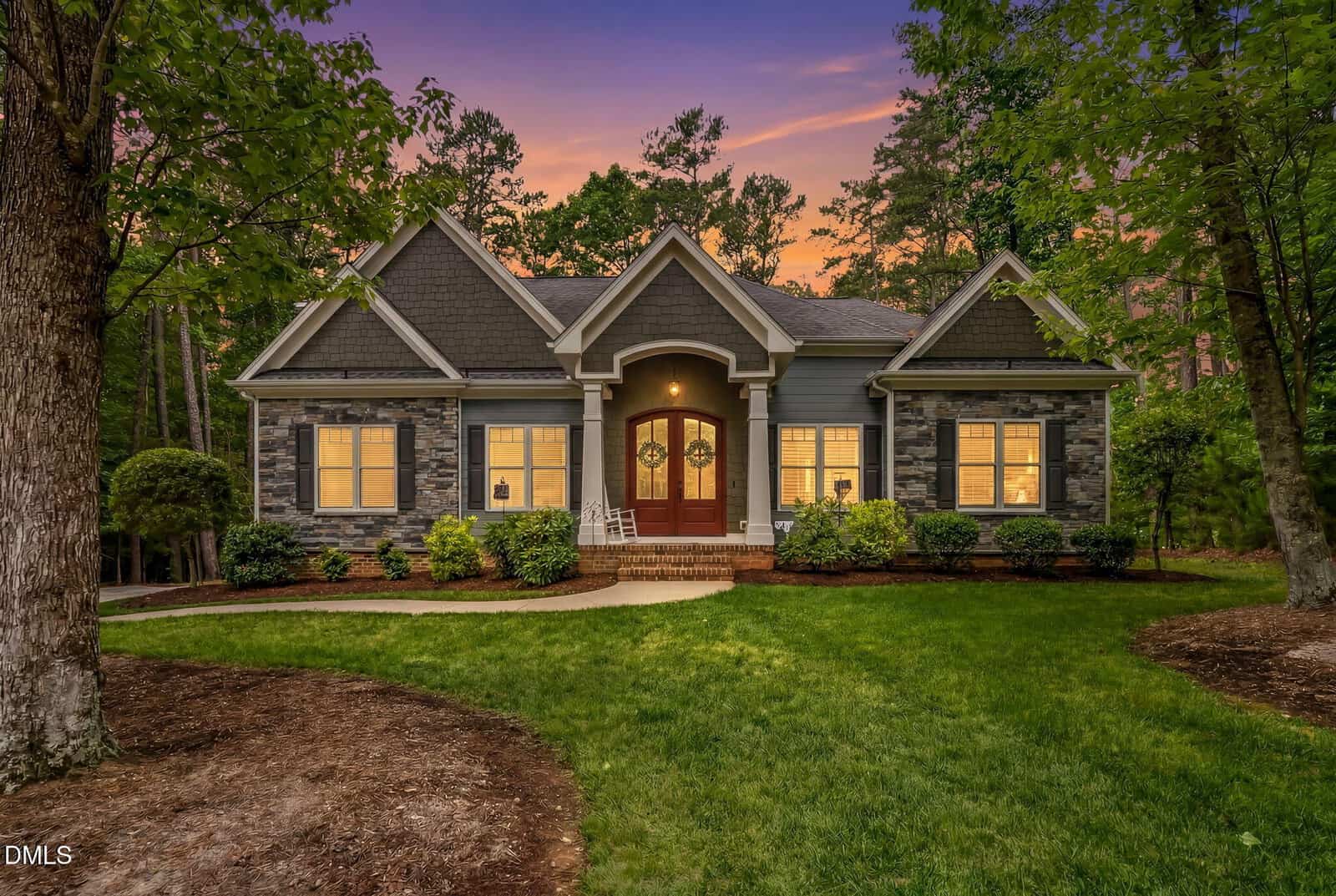 Single-story house with stone and gray siding, warmly lit windows, double wooden front doors, front porch with plants, and a well-manicured lawn surrounded by tall trees at sunset.