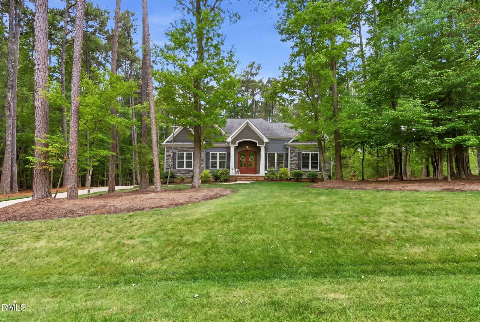 A single-story house with gray siding and white trim sits surrounded by tall trees. The front lawn is large and well-manicured, and a curved sidewalk leads to the front porch with double doors.