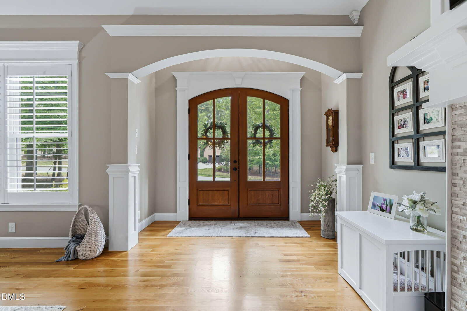 A bright entryway with double wooden doors featuring glass panes, flanked by white trim and decorative columns. Hardwood floors, a basket with a blanket, and a side table with a vase and framed photos are visible.