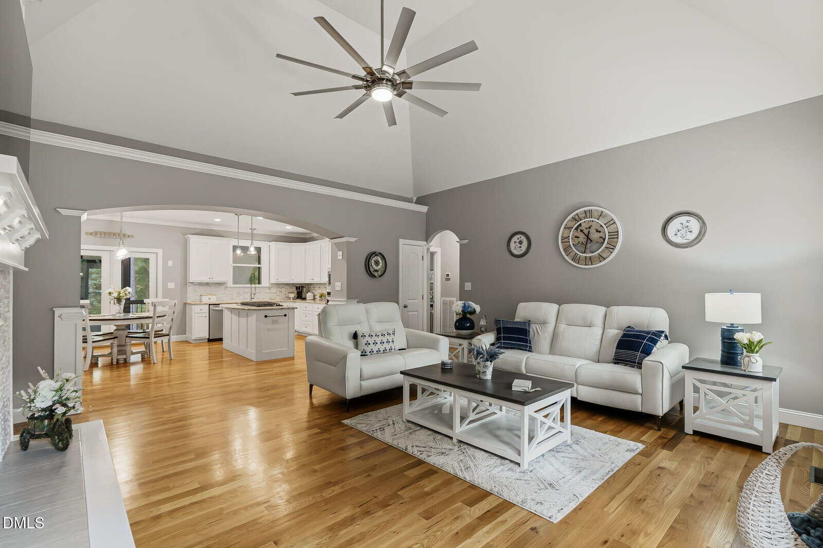 Modern living room with white sofas, a coffee table, decorative clocks on gray walls, hardwood floors, and an open view into a white kitchen and dining area, all under a high ceiling with a large ceiling fan.