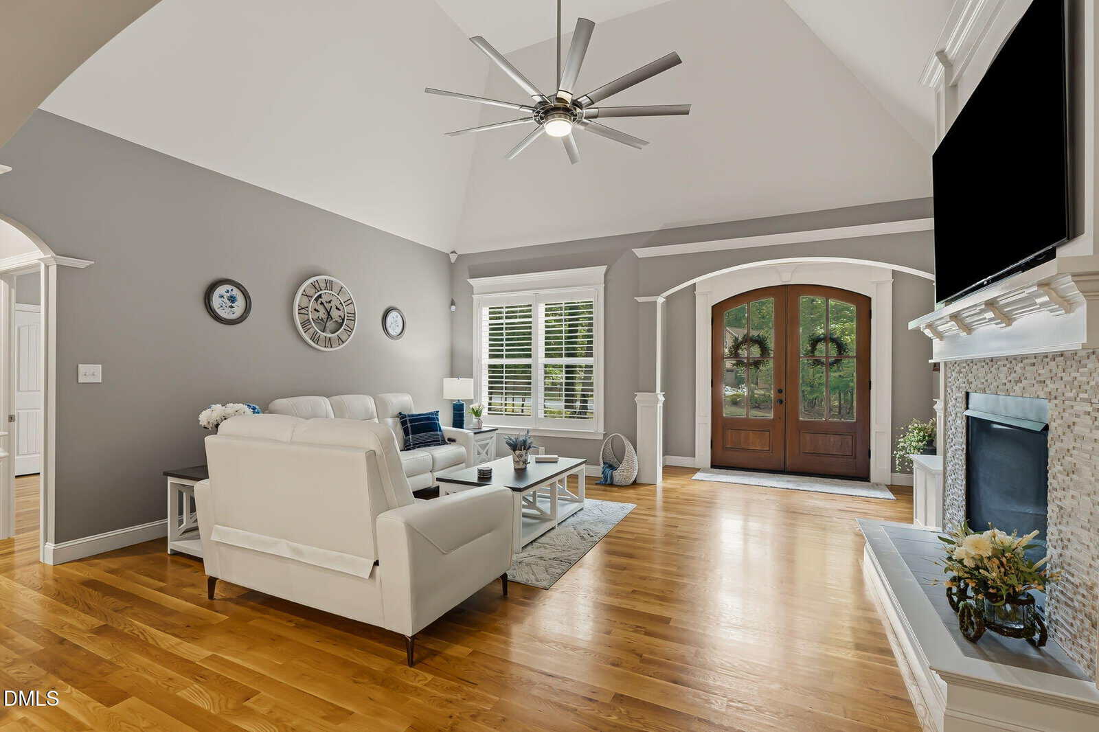 Modern living room with light gray walls, vaulted ceiling, hardwood floors, white furniture, a fireplace, wall-mounted TV, large windows, and double wooden front doors letting in natural light.