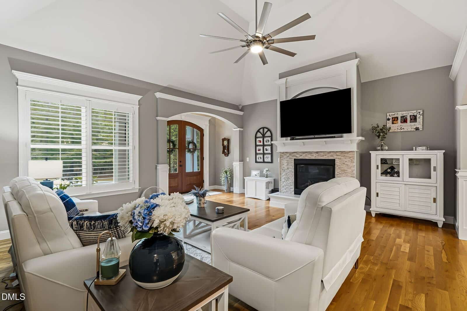 Bright living room with white furniture, hardwood floors, a large window with blinds, a dark wood front door, a fireplace with a TV above it, and modern decor, including blue and white floral arrangements.